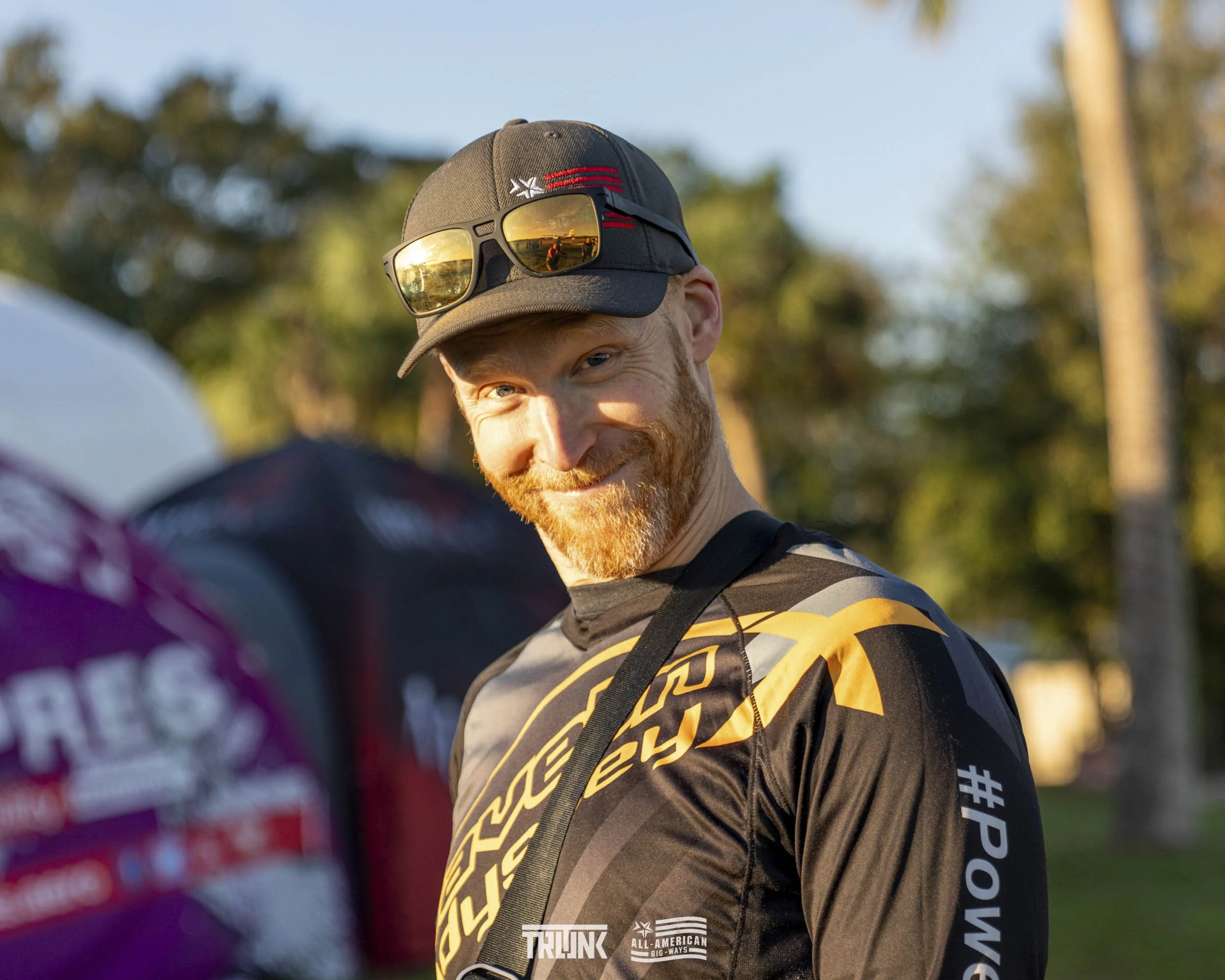 Smiling man with a beard wearing a black cap with sunglasses on top, outdoors during sunset, in a black and yellow sports jersey, with trees and blue sky in the background.