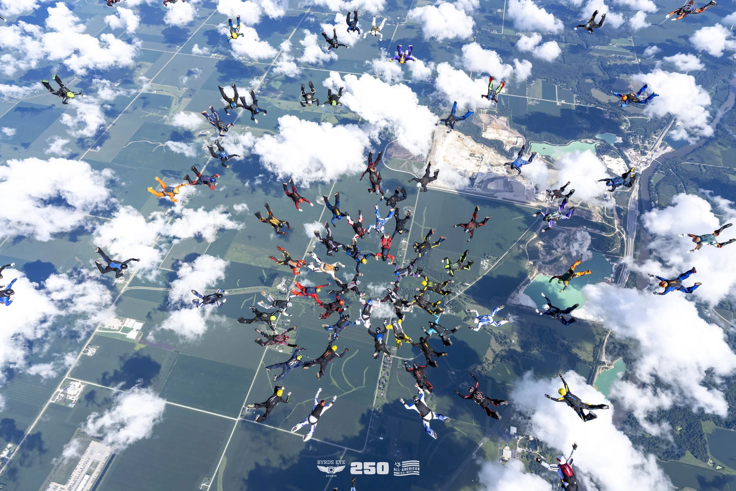 Aerial view of a large group of skydivers in colorful jumpsuits free-falling over a landscape with fields, roads, and lakes, surrounded by clouds.