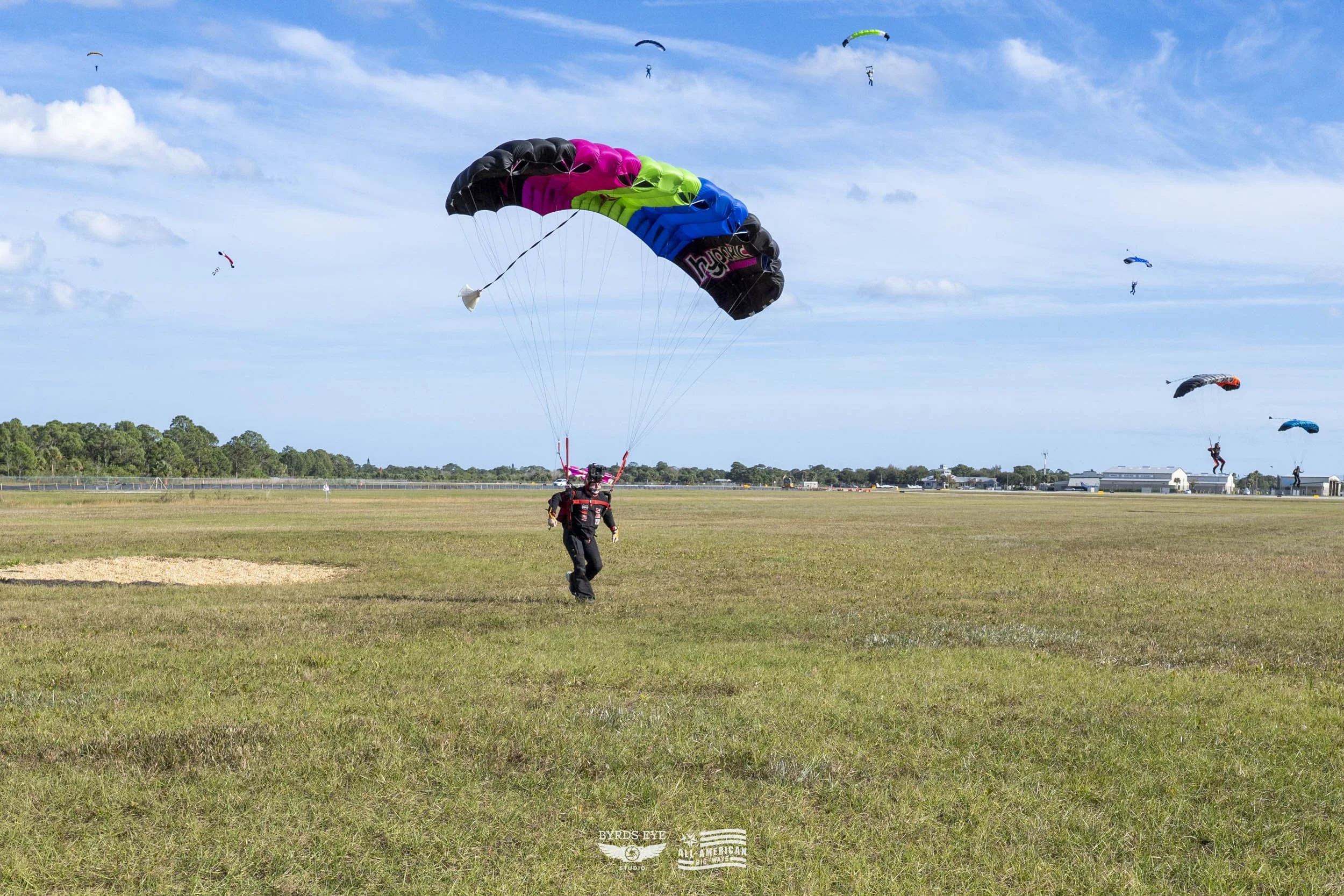 A person on the ground preparing to skydive with a colorful parachute, while several other skydivers descend with their parachutes in the sky over an open grassy field under a partly cloudy sky.