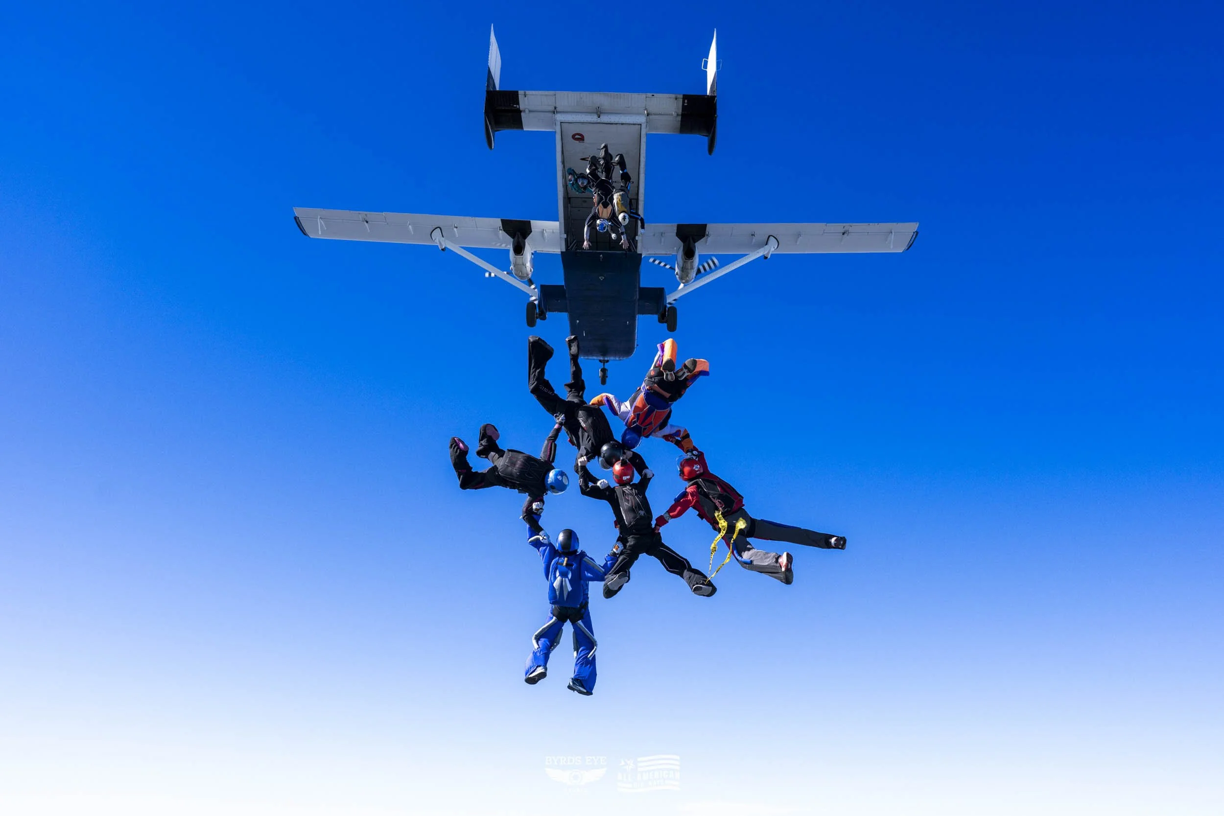 Skydivers in a freefall formation beneath an airplane against a blue sky.