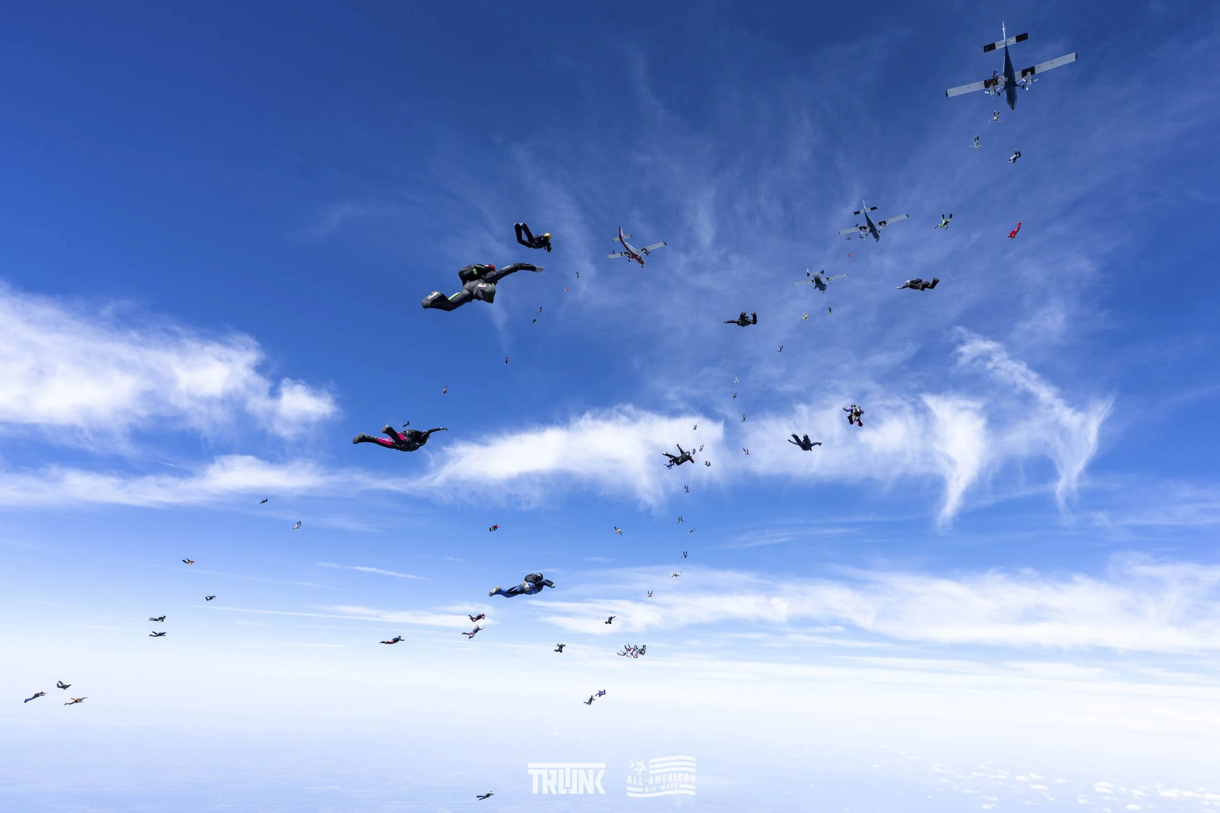 Skydivers in free fall formation against a blue sky with scattered clouds, some with parachutes deployed, flying in various positions and orientations.