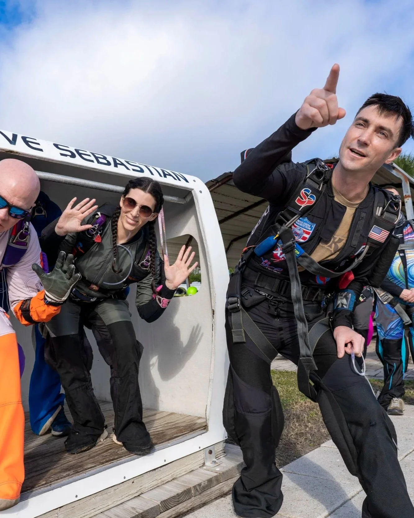 Skydivers in jumpsuits and gear posing outside an aircraft, with some waving and one person pointing upward.