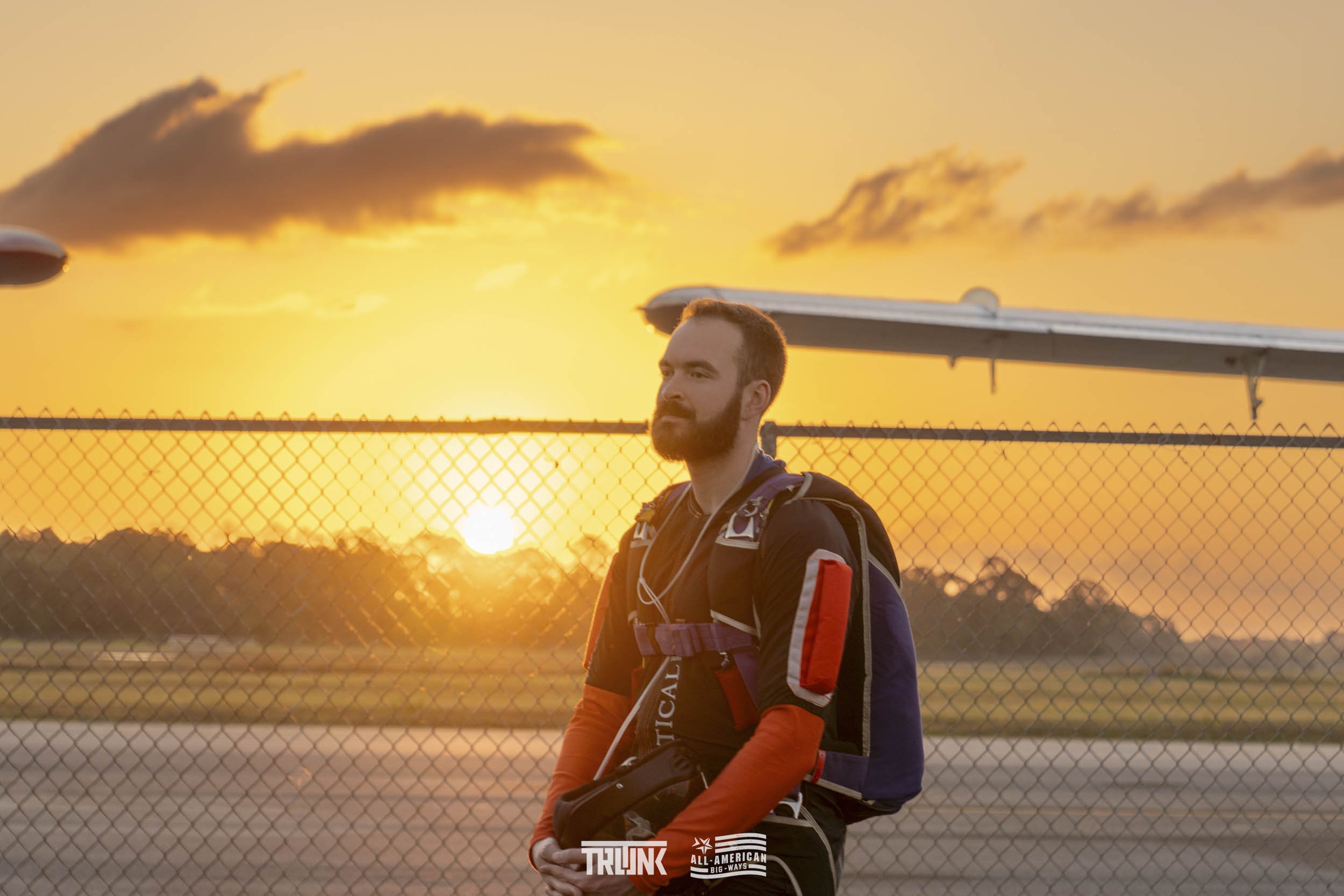 A man standing outside at sunset with a backpack over his shoulders, wearing a black jacket with red accents, near a chain-link fence and an airplane in the background.