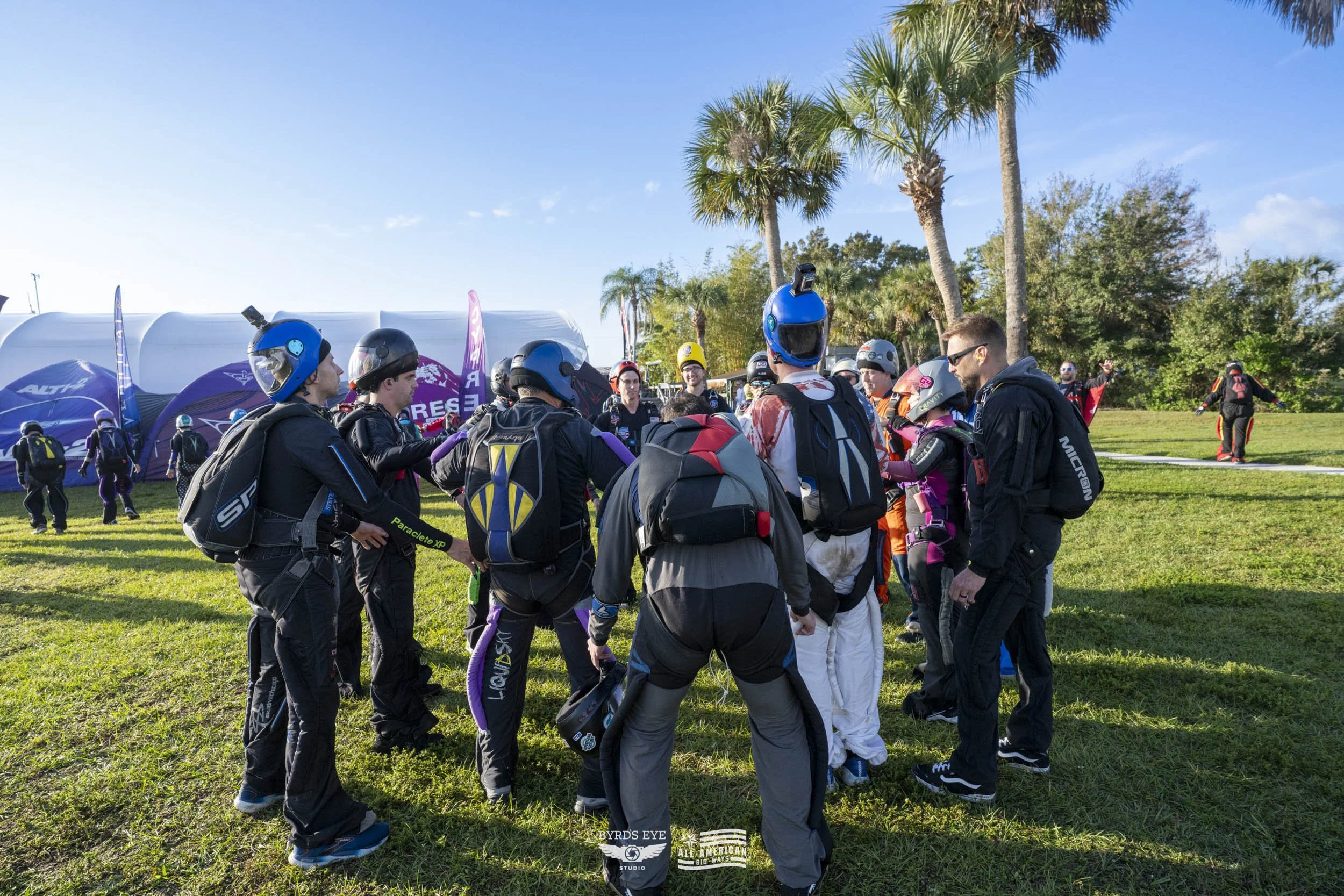Group of skydivers gathered outdoors, wearing jumpsuits, helmets, and backpacks, preparing for a skydiving session on a sunny day with palm trees in the background.