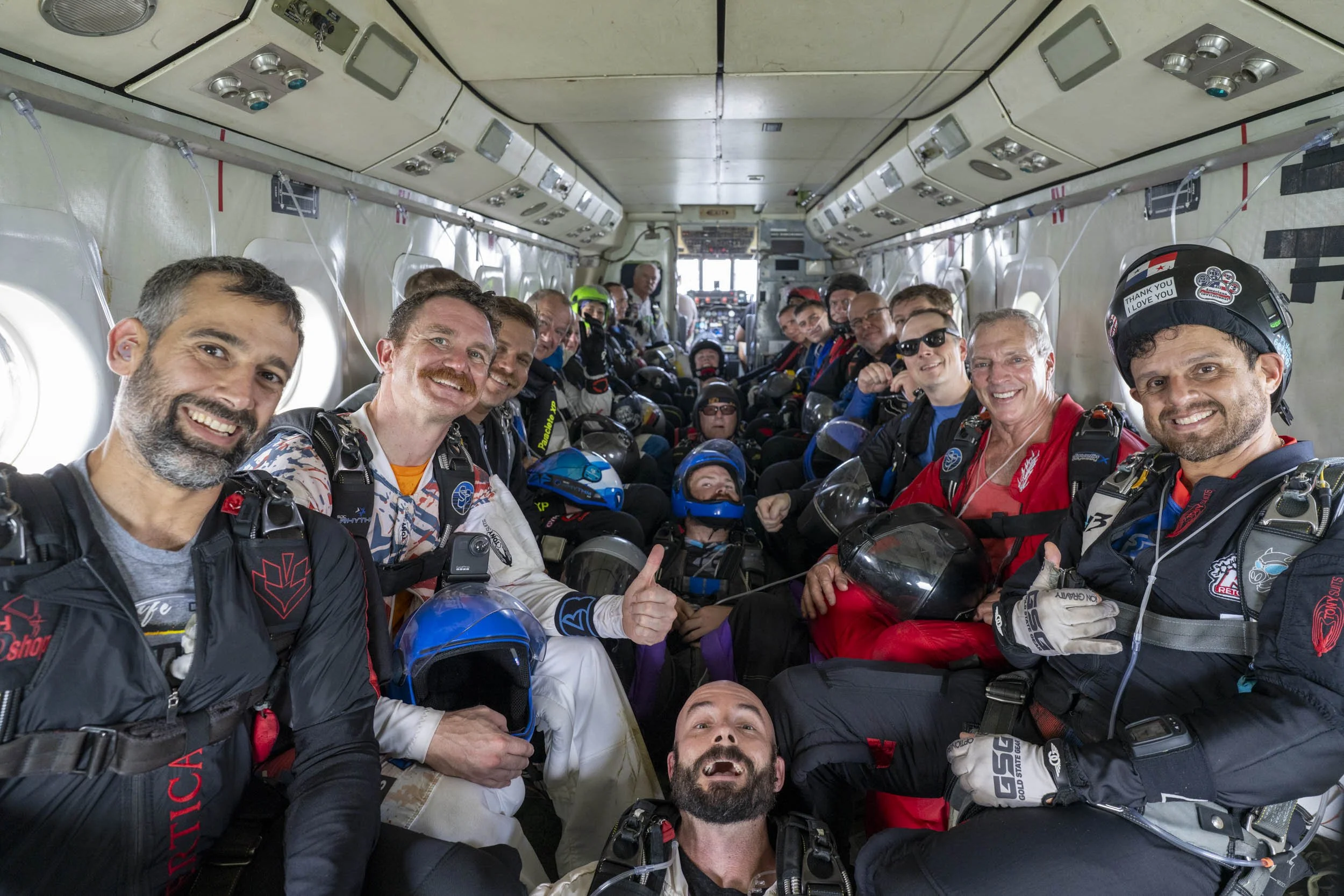 A group of skydivers sitting inside an aircraft, smiling at the camera, wearing jumpsuits and helmets, with their helmets and gear in their laps and on their laps.