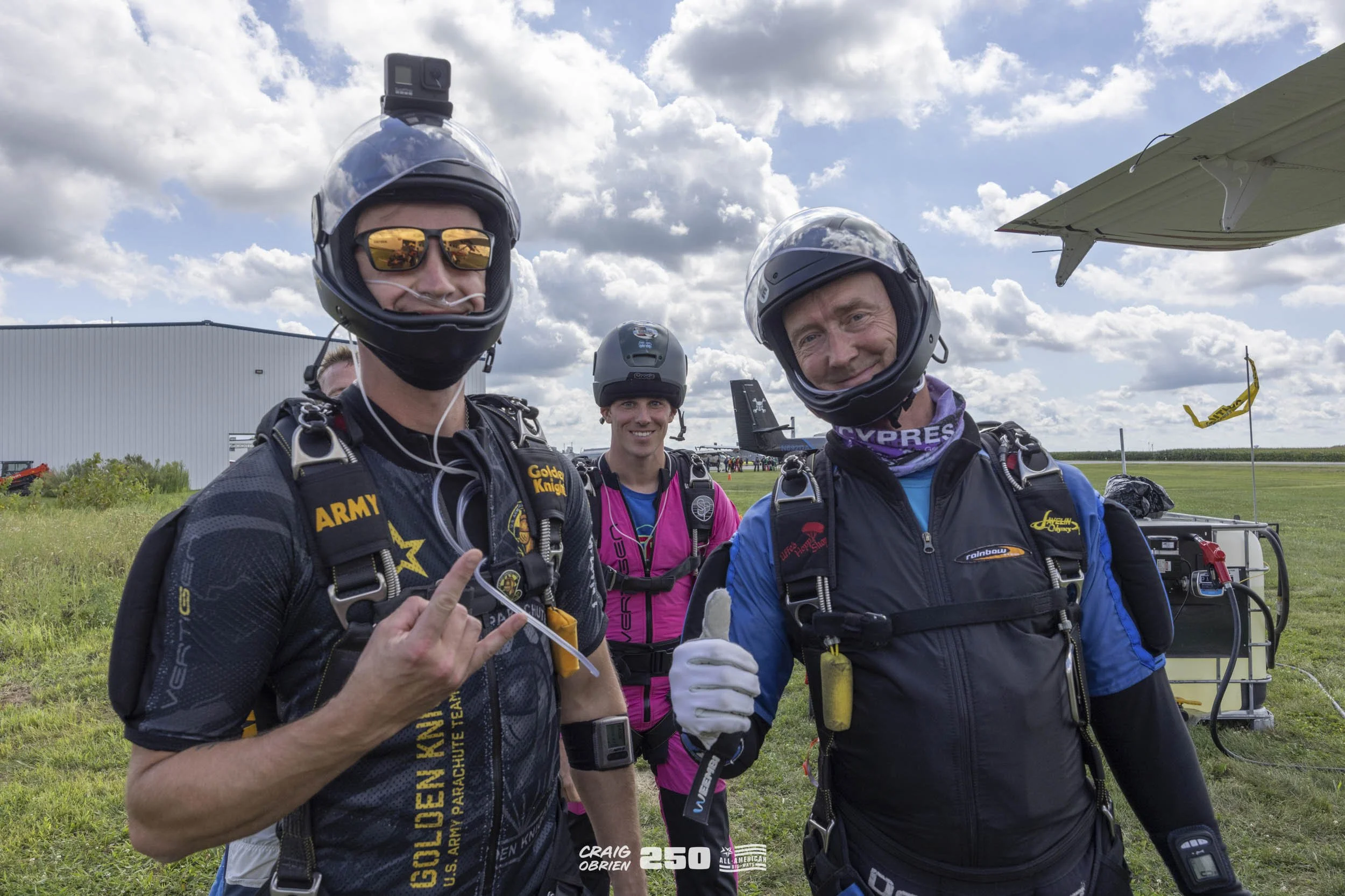 Three skydivers in jumpsuits and helmets standing on grass, smiling for the camera, with a small aircraft and cloudy sky in the background.