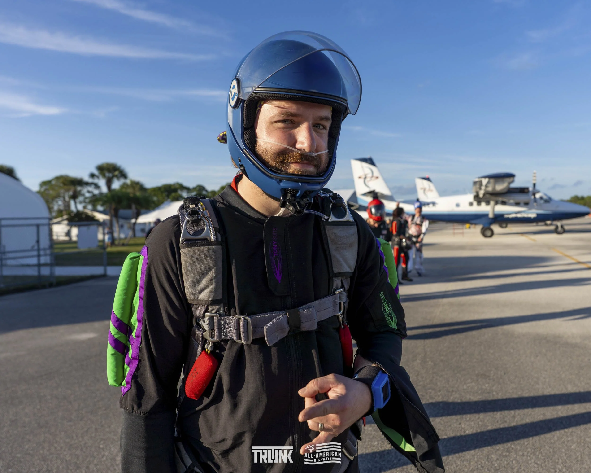 A man in skydiving gear standing outdoors on a sunny day, with airplanes and other skydivers in the background.