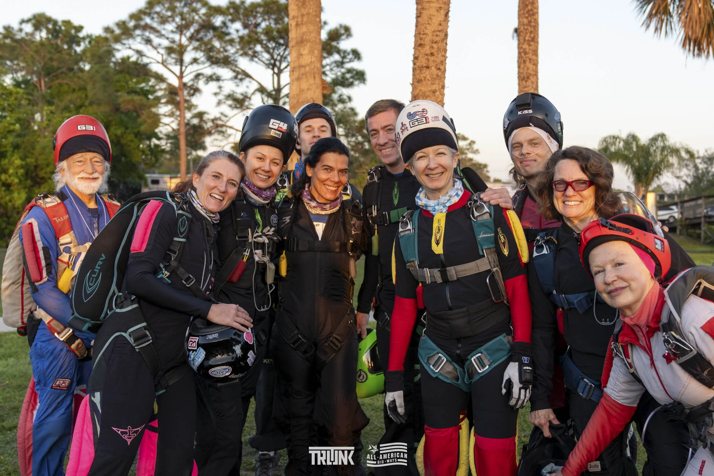 Group of skydivers in jumpsuits and helmets standing outdoors with trees in the background, smiling at the camera.