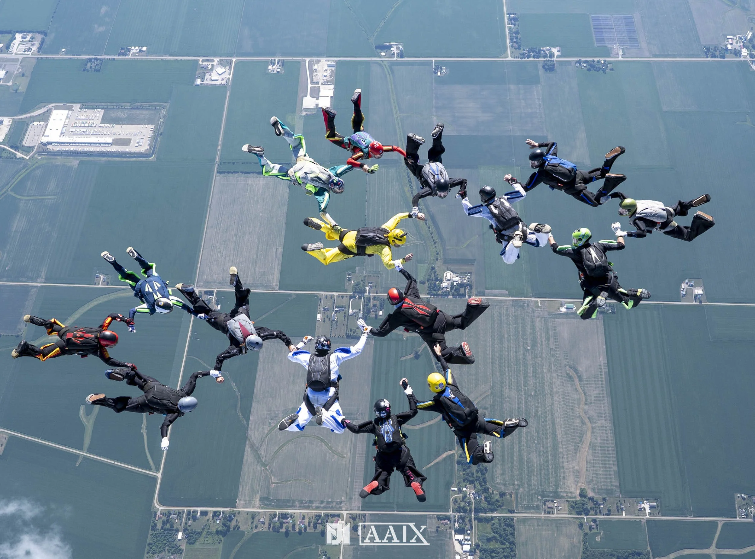 Skydivers forming a circle in freefall over farmland and fields, wearing colorful jumpsuits and helmets.