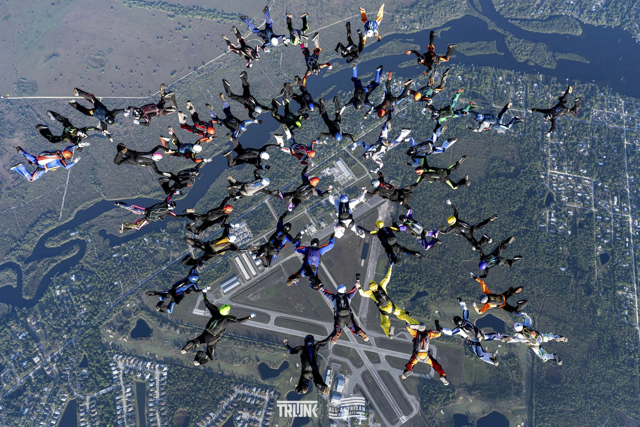 Group of skydivers in free fall over a landscape with rivers, roads, and buildings.