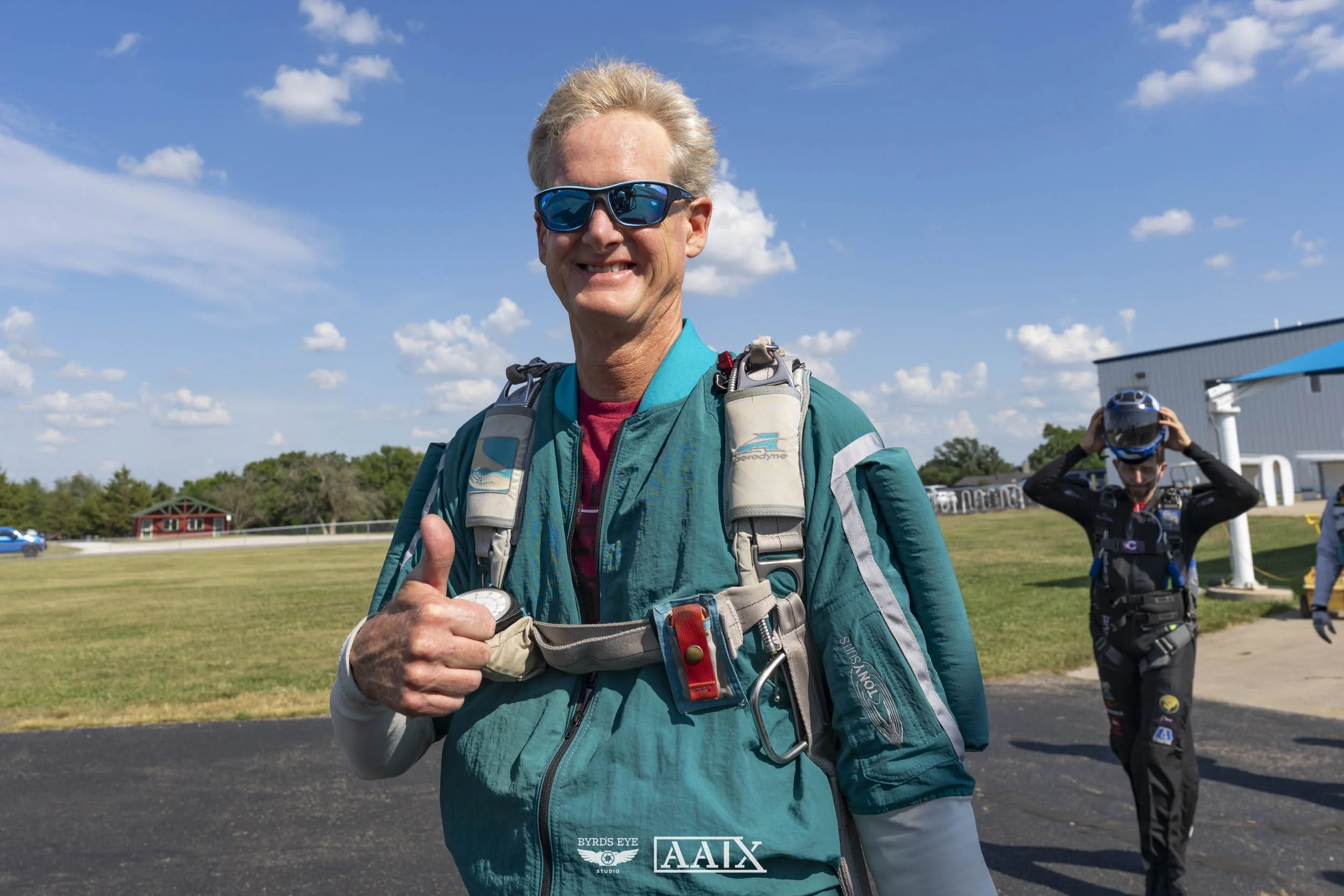 An older man with blonde hair wearing sunglasses, a teal jacket, and a parachute harness, giving a thumbs-up outdoors on a sunny day, with a younger man in skydiving gear and helmet in the background.