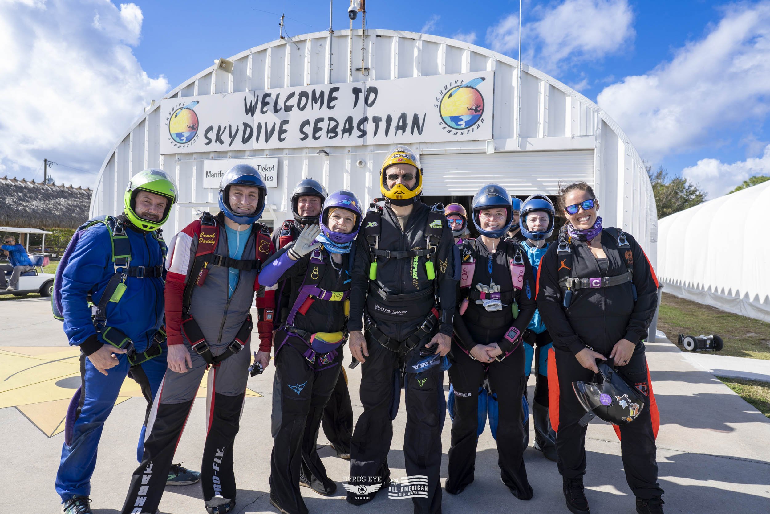 Group of nine skydivers in jumpsuits and helmets standing in front of Skydive Sebastian entrance, smiling and posing for the photo.