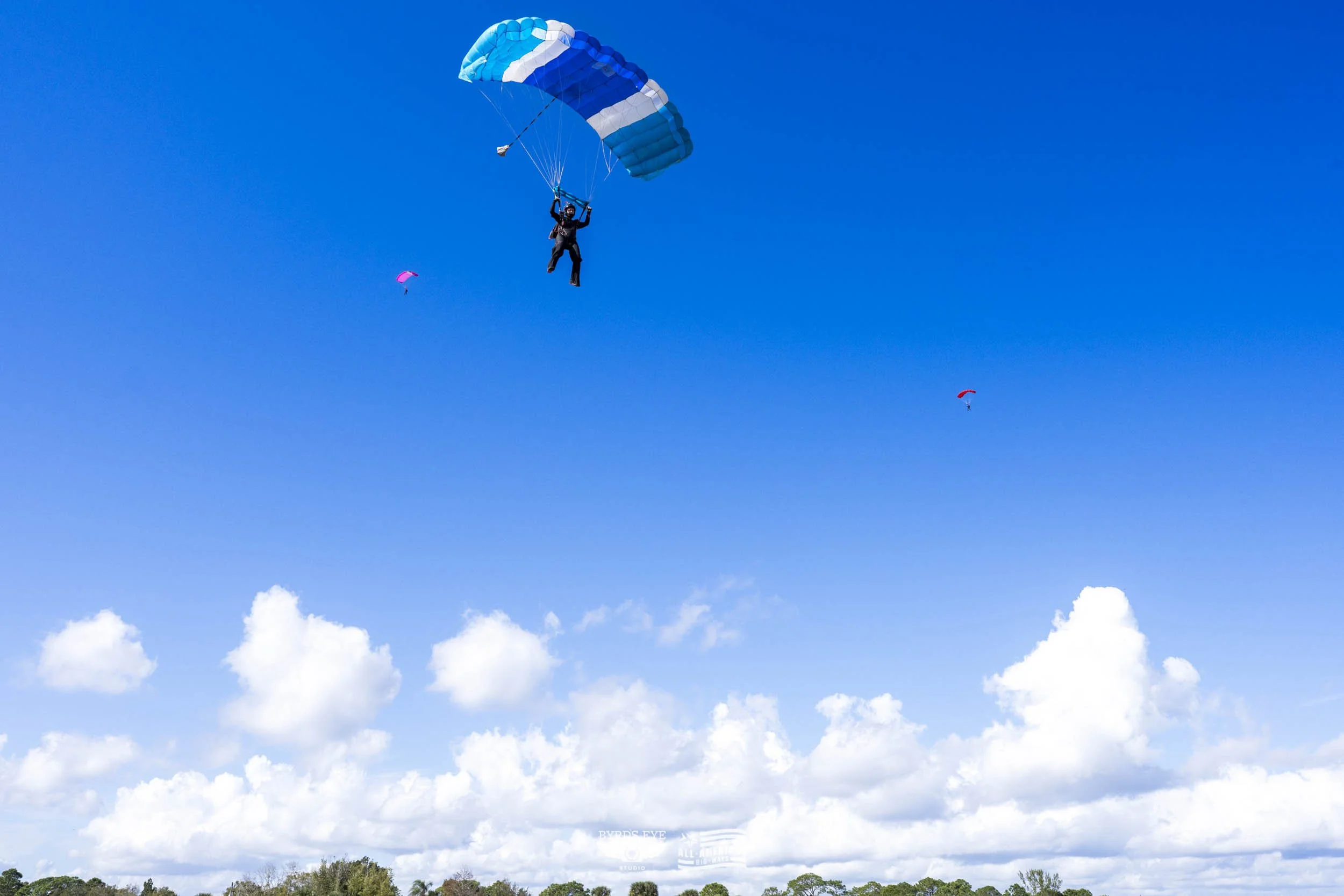 A person skydiving with a blue and white parachute, falling through a bright blue sky with scattered white clouds and two smaller parachuters in the distance.