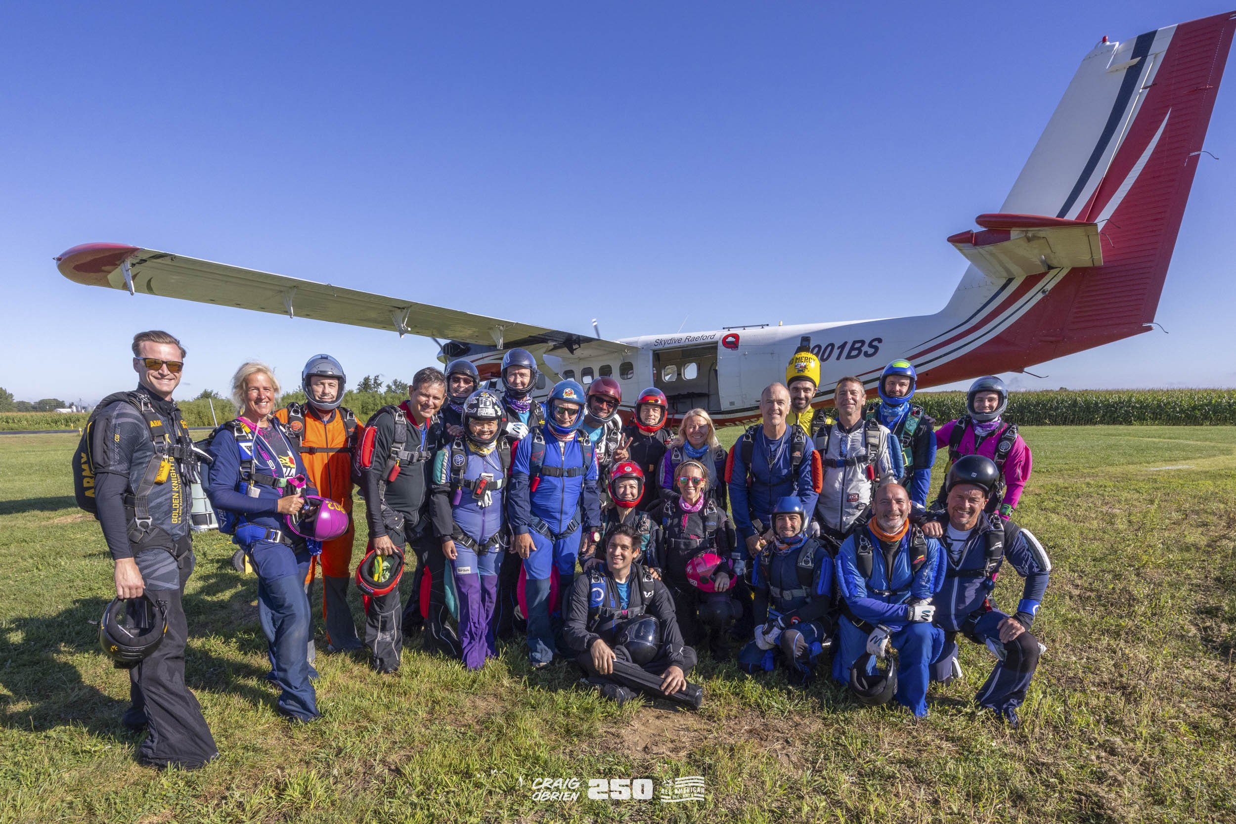 Group of skydivers in jumpsuits and helmets standing on grass in front of a small airplane on a clear, sunny day.
