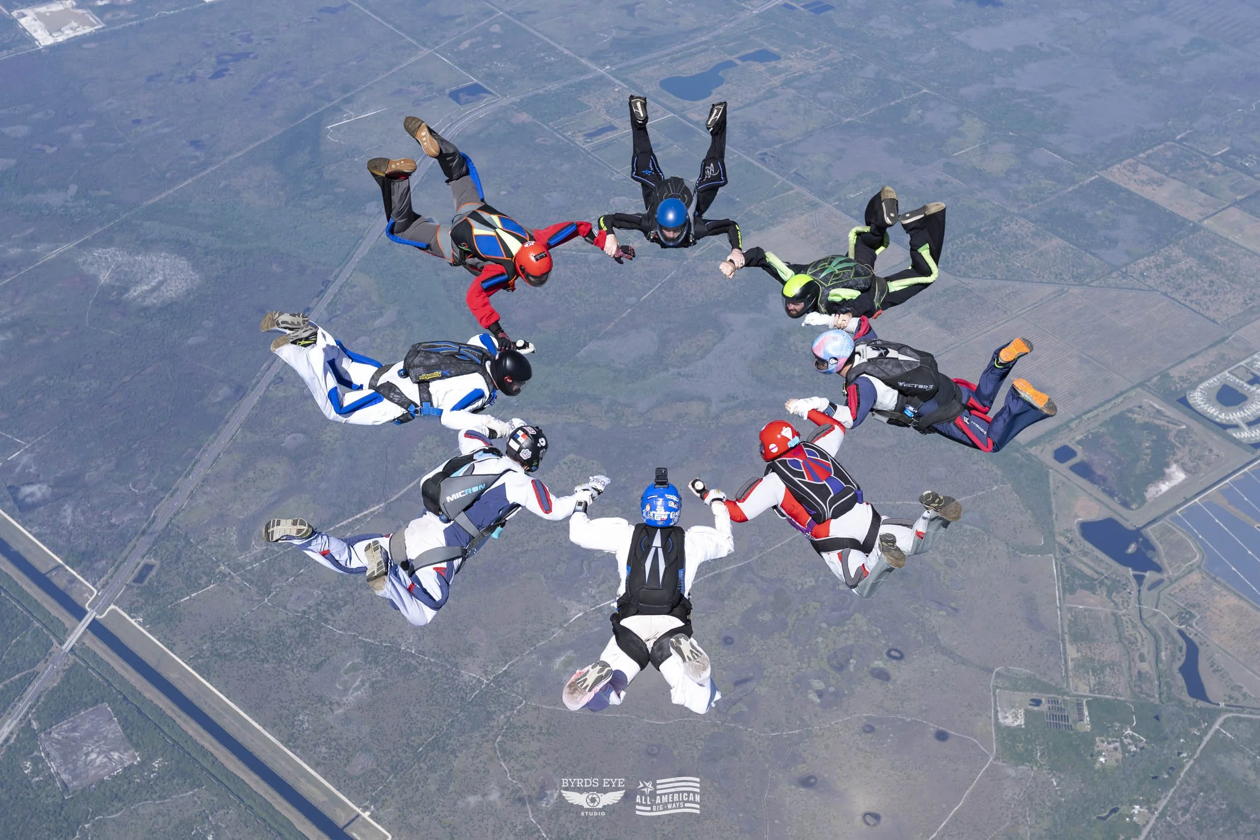 A group of skydivers holding hands and forming a circle while free-flying in mid-air over a landscape with fields, roads, and a water body.