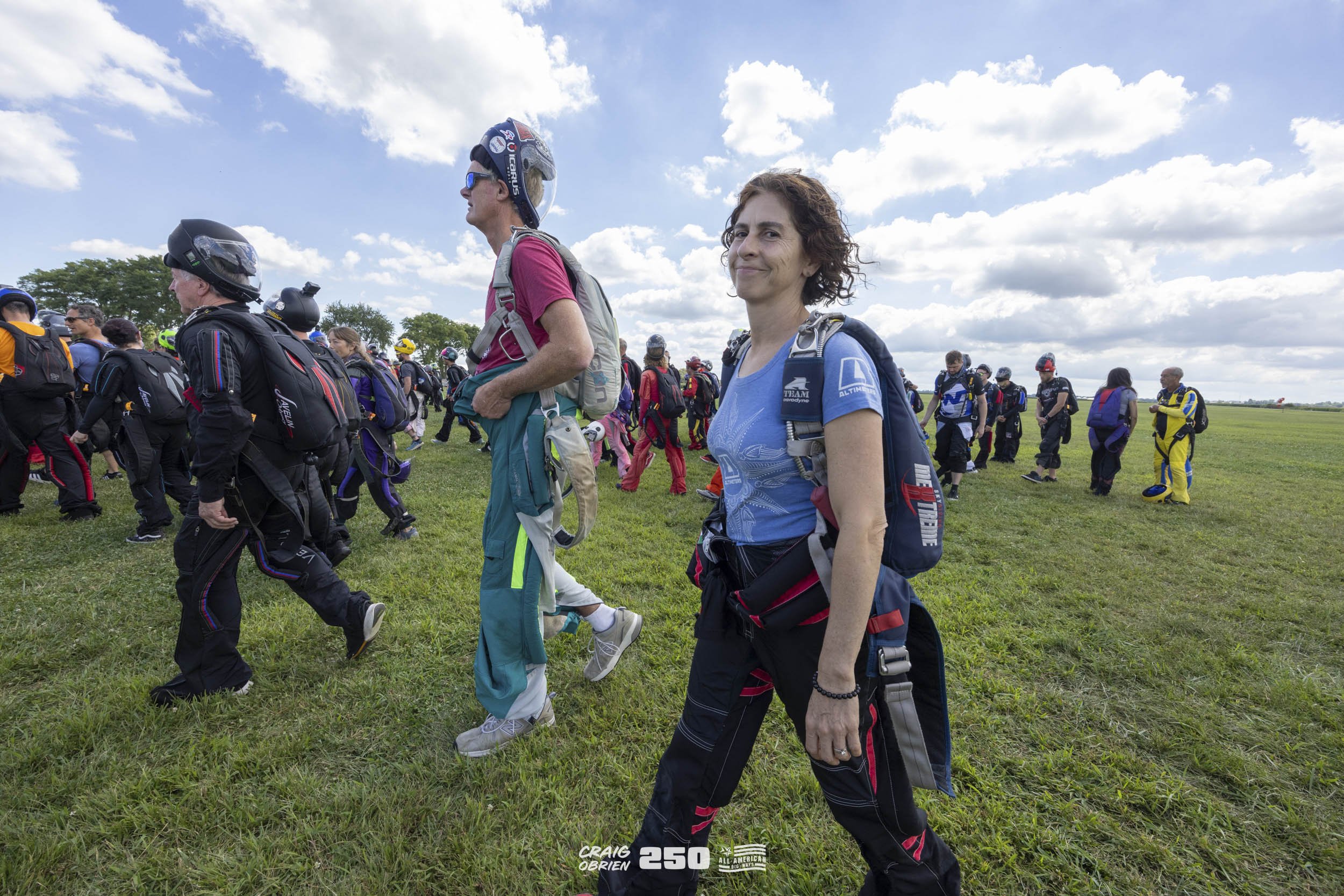 Group of skydivers on a grassy field preparing for a jump, with some wearing helmets and harnesses, under a partly cloudy sky.