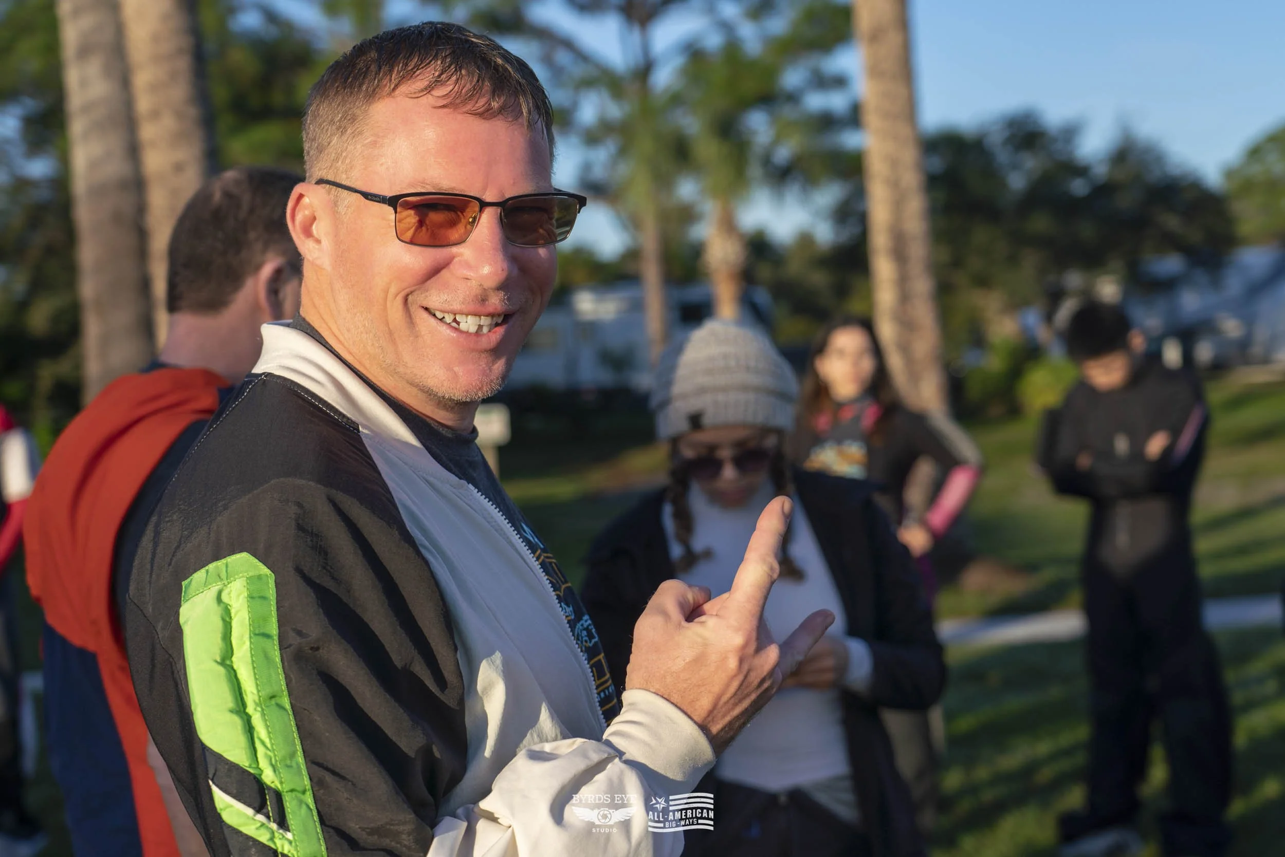 A smiling man wearing sunglasses and a racing jacket makes a rock and roll hand gesture at an outdoor gathering, with other people and trees visible in the background.