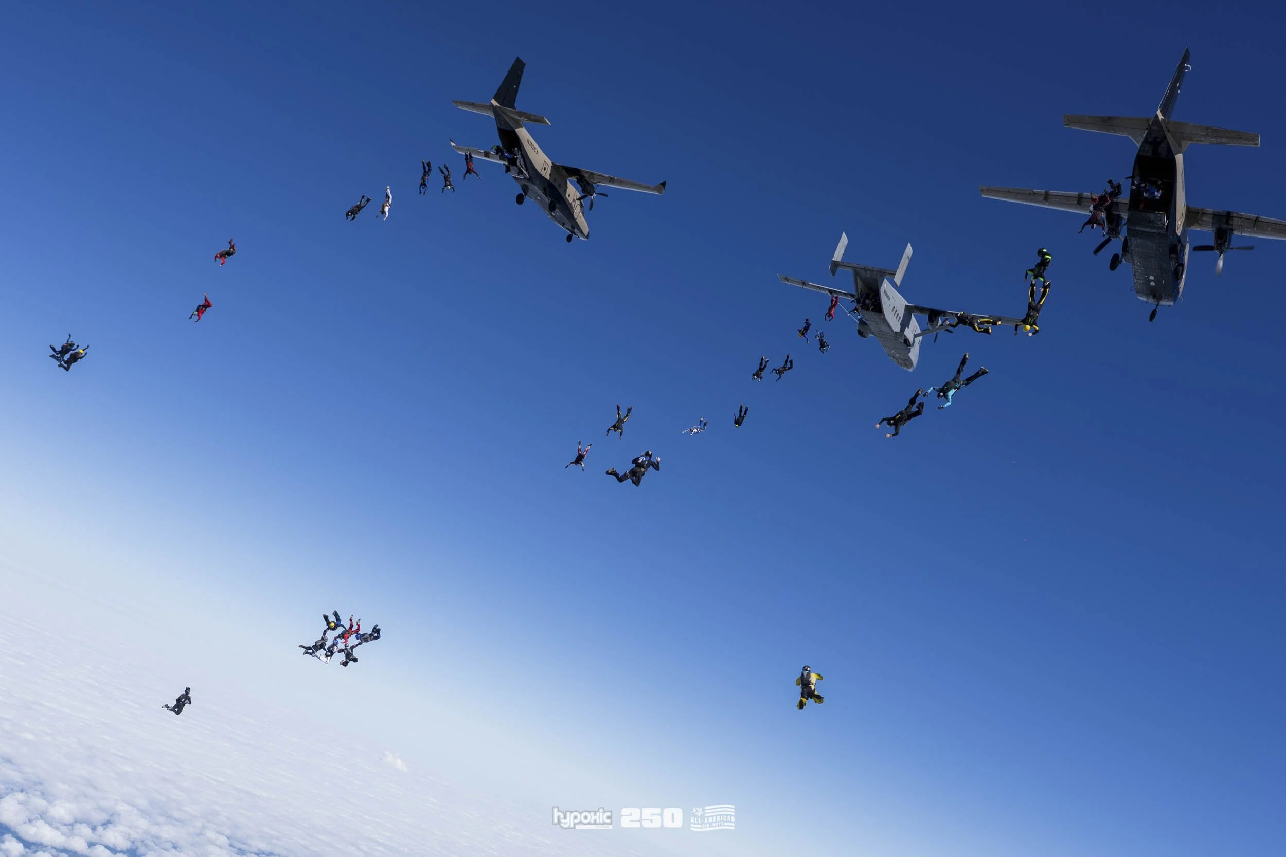 Skydivers and airplanes in mid-air during a jump, against a clear blue sky with clouds below.