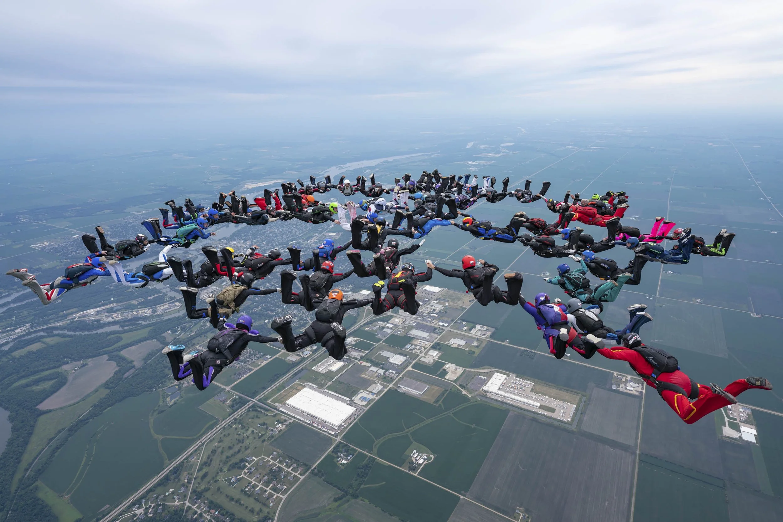Group of skydivers in a synchronized formation during freefall over a landscape with fields and buildings.