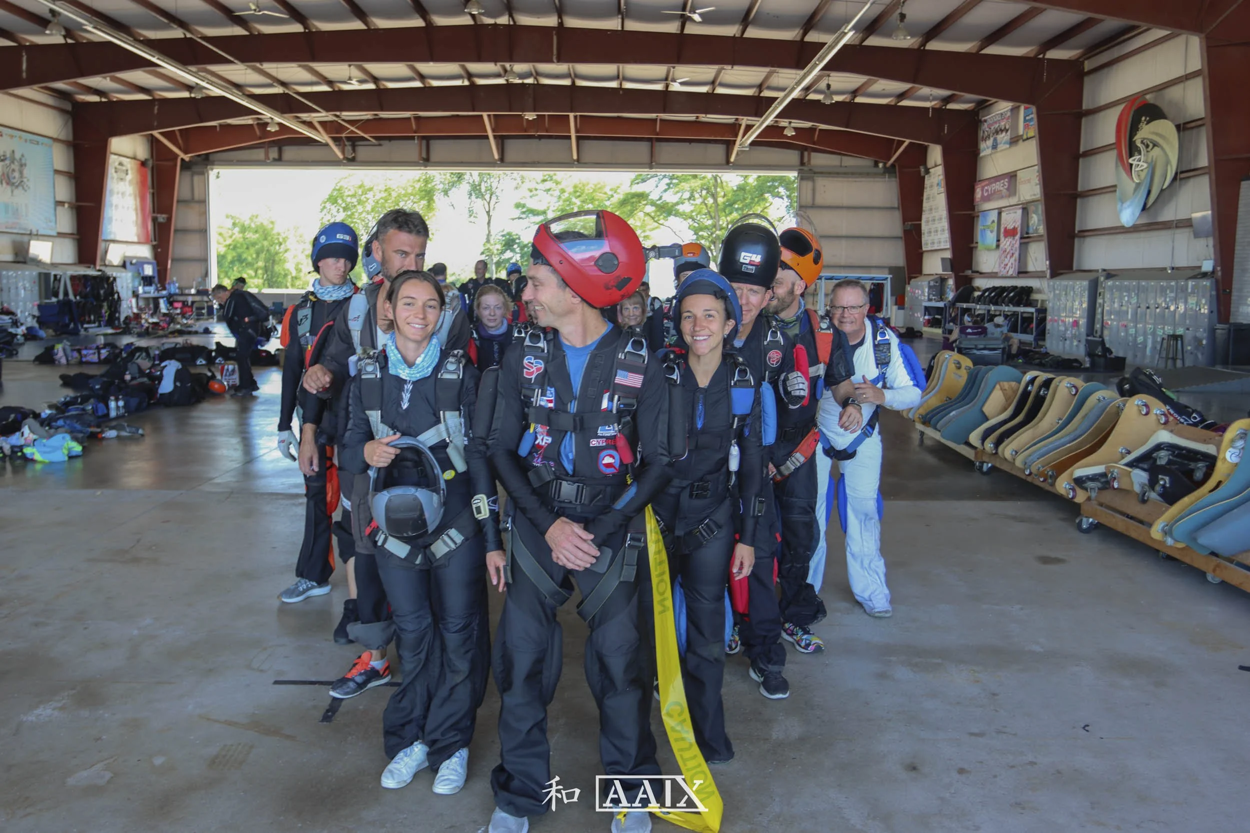 Group of skydivers in jumpsuits and helmets standing inside a hangar with parachutes and gear around them, smiling and preparing for a jump.