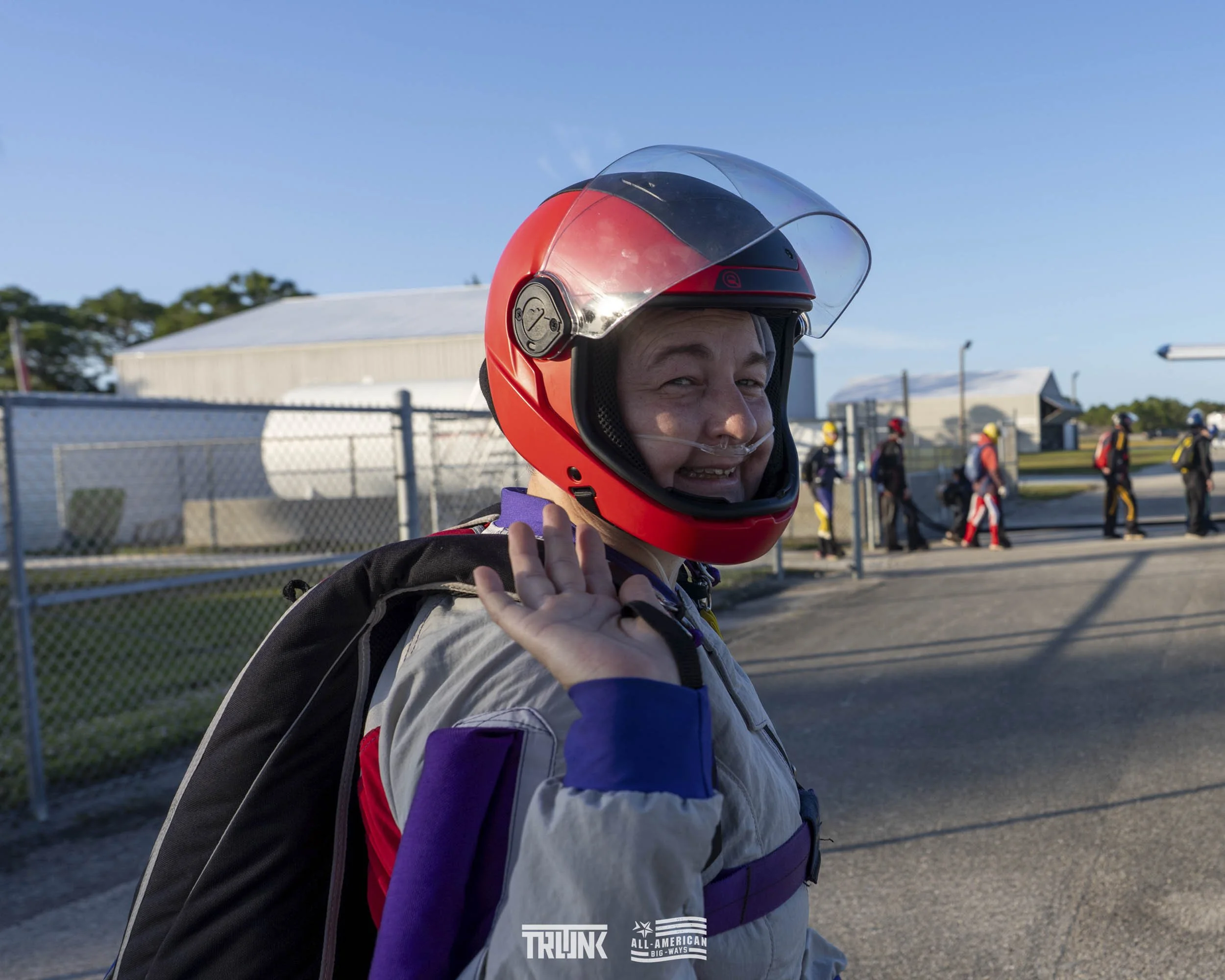 Person in racing gear and a red helmet waving at the camera, with a group of people in racing suits walking on the background at a race track.
