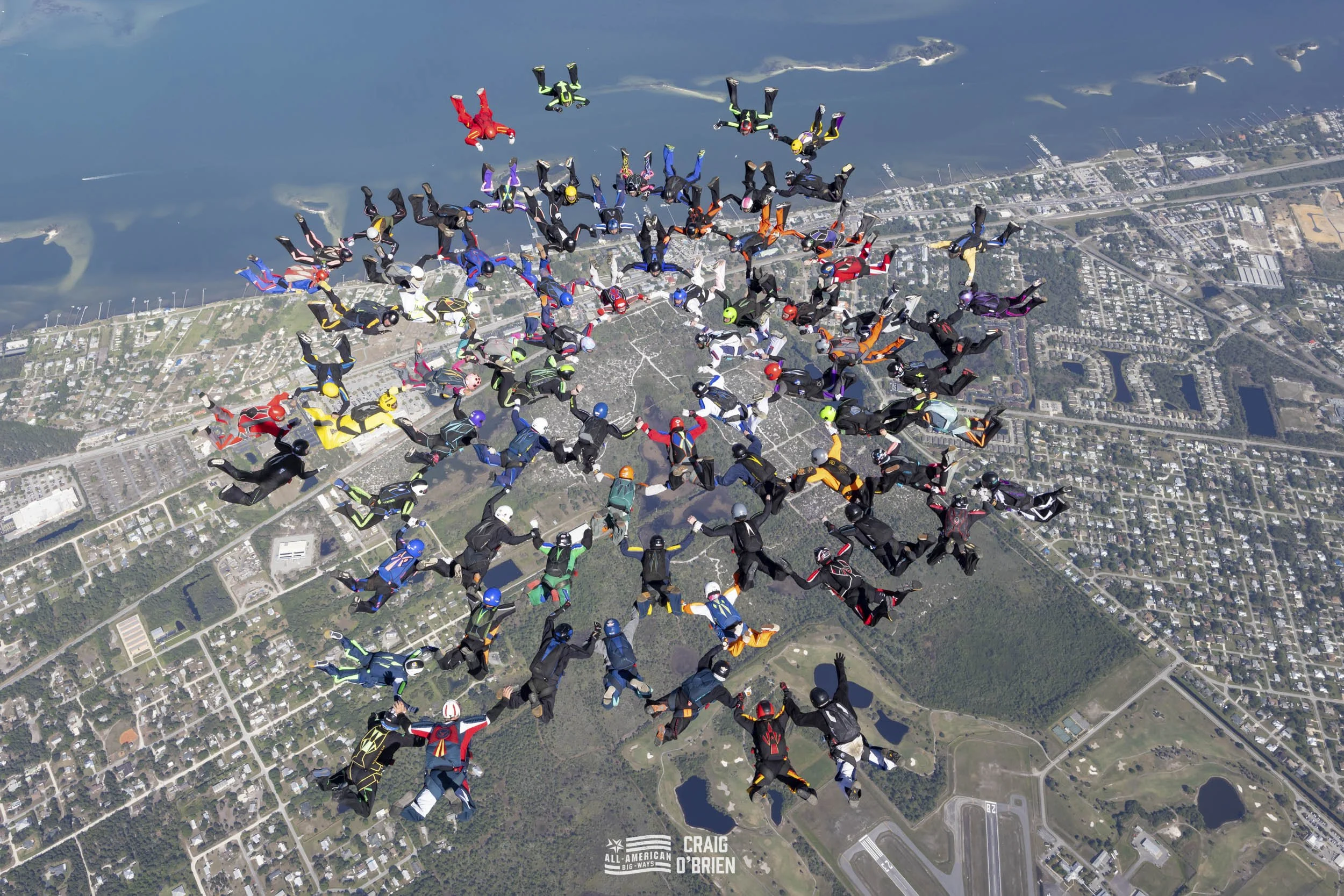 A large formation of skydivers in colorful jumpsuits and helmets free-falling in a circular pattern over a coastal city with water and land visible below.