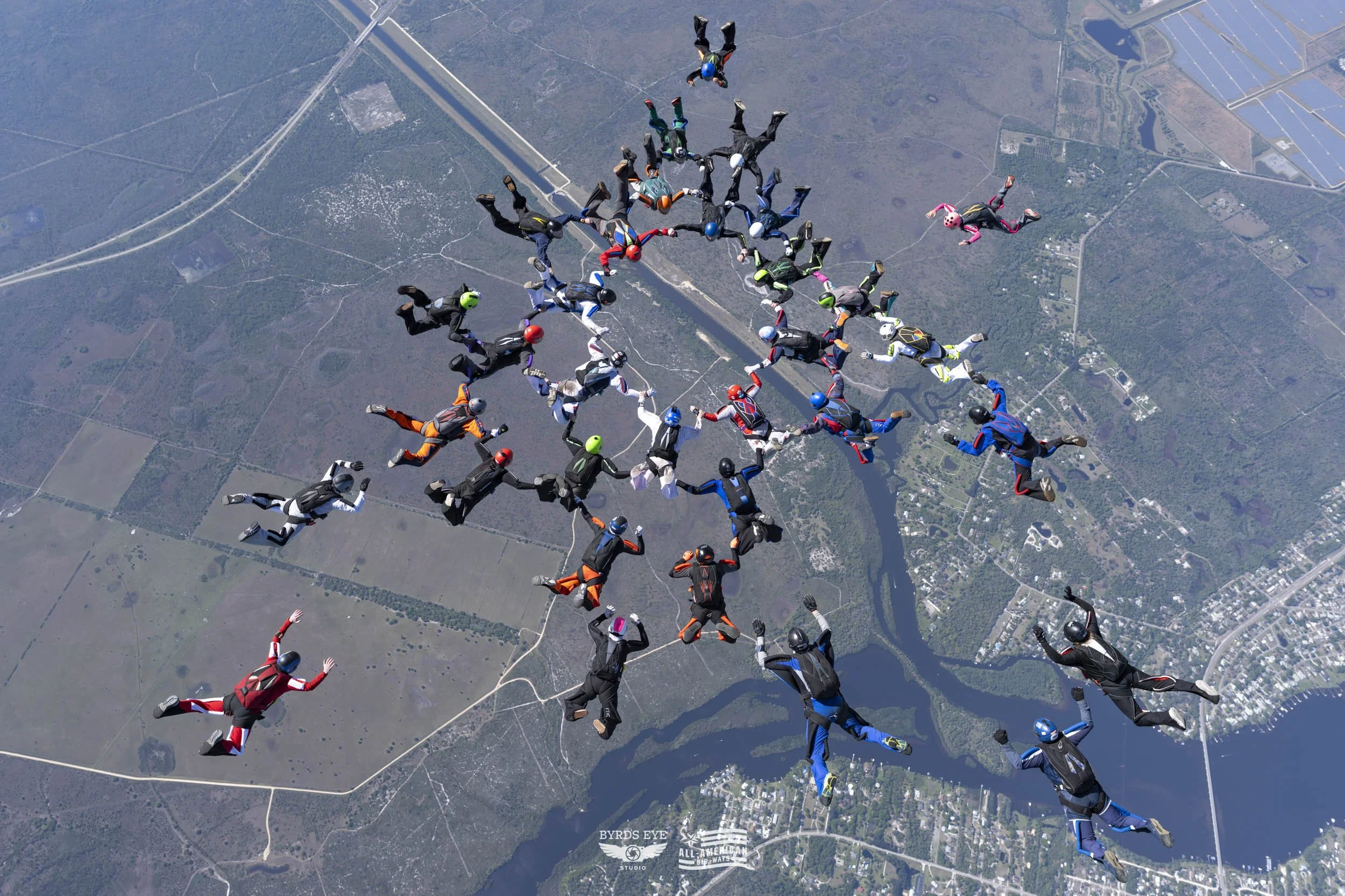 Skydivers in colorful jumpsuits and helmets forming a star shape in free fall above a landscape with roads, rivers, and fields.