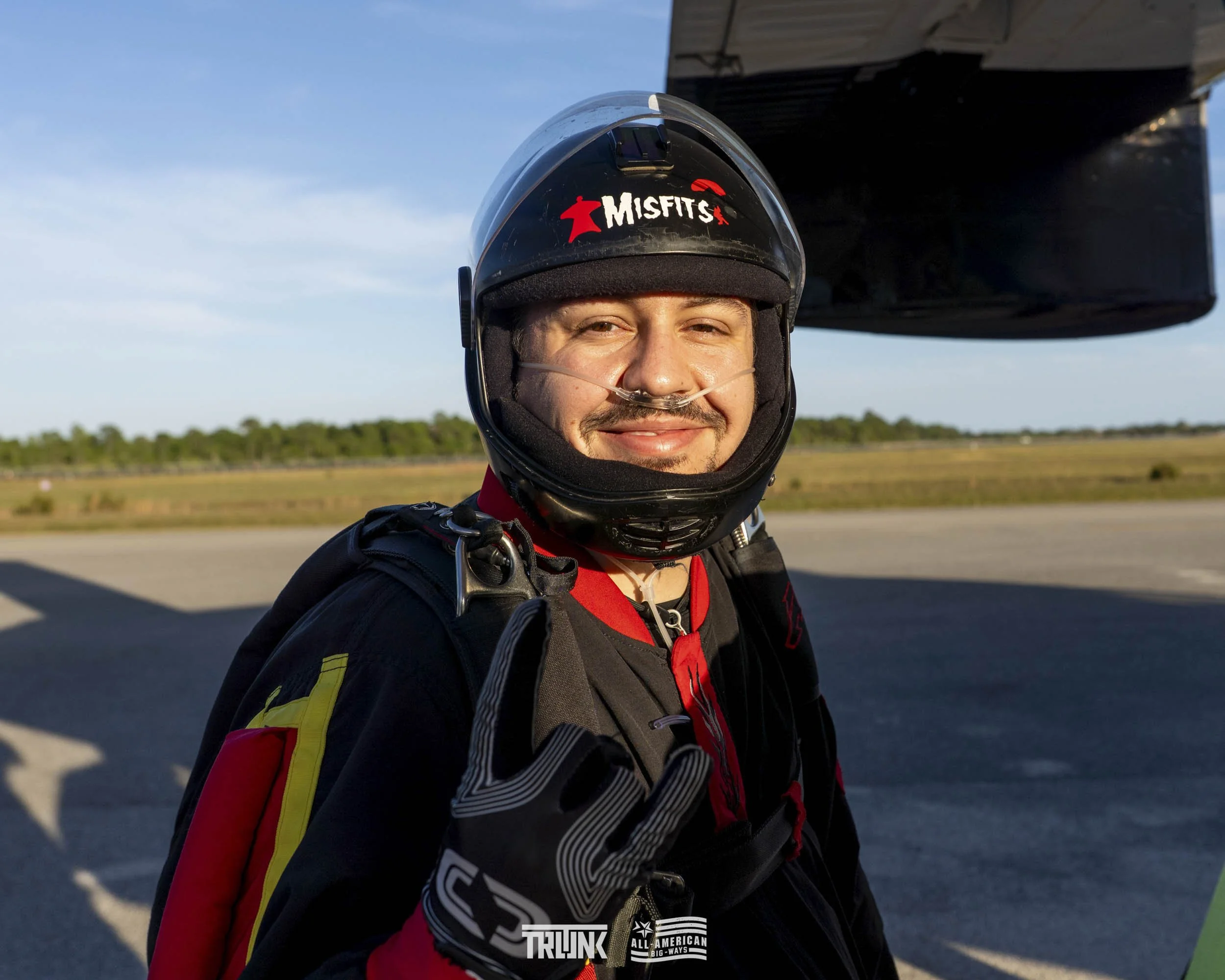 A smiling male skydiver wearing a black helmet with a red Misfits logo, a breathing tube, and a black jumpsuit with red and yellow accents, standing on the tarmac with open sky and flat landscape in the background.
