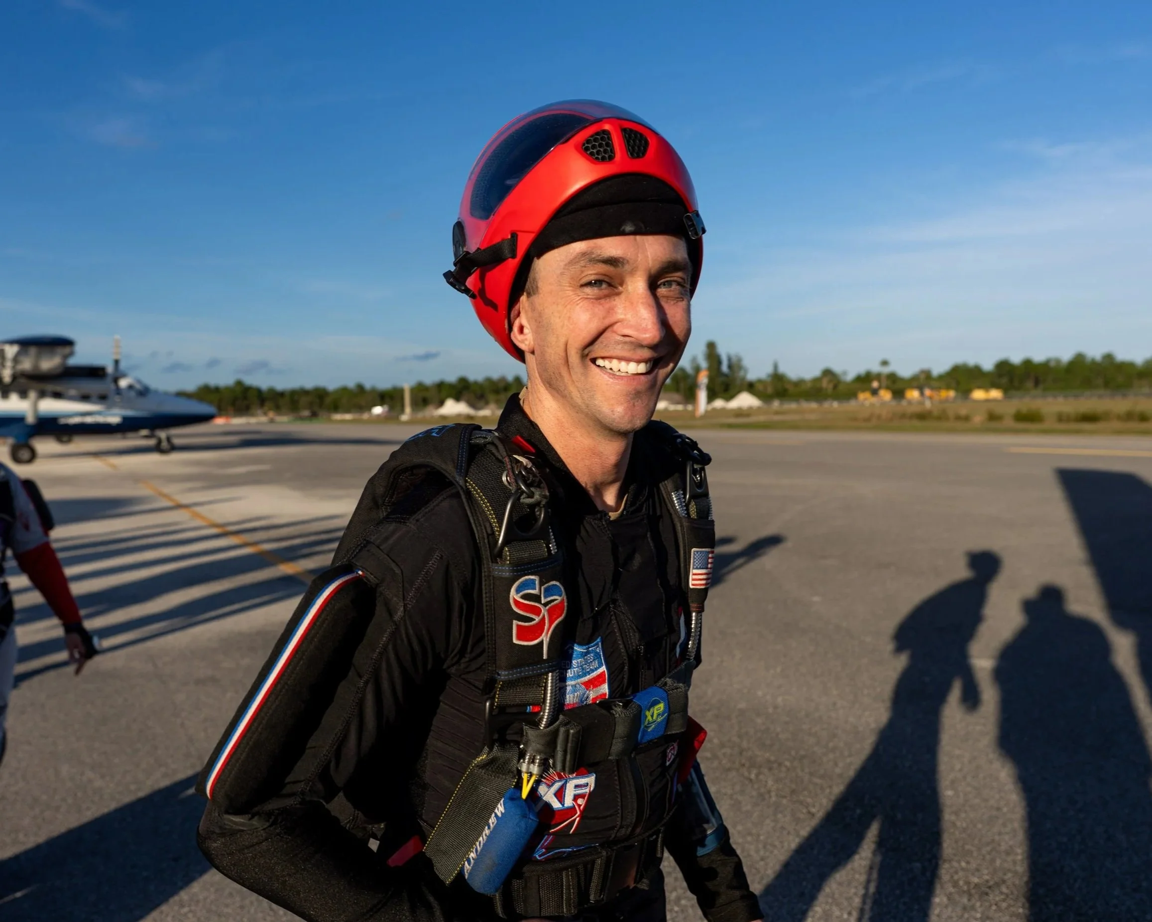 A smiling man in a skydiving suit and red helmet stands on an airport tarmac.