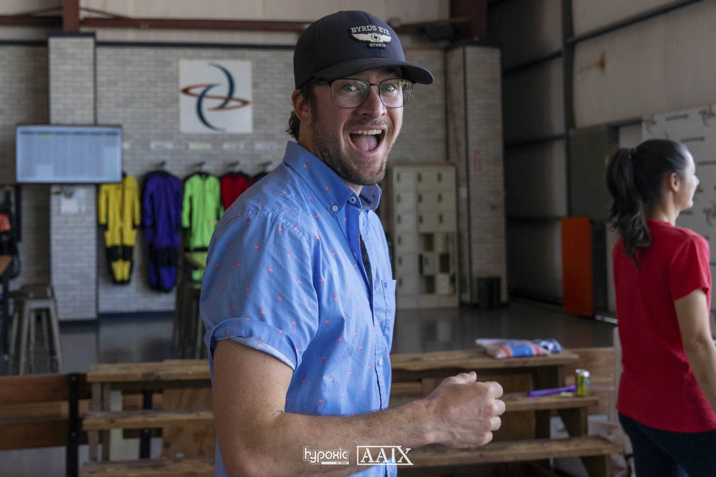 A man with glasses, a beard, and a mustache wearing a baseball cap and a blue shirt with pink flamingos, smiling and looking at the camera inside a bowling alley or recreational space with colorful jackets hanging on a wall in the background.