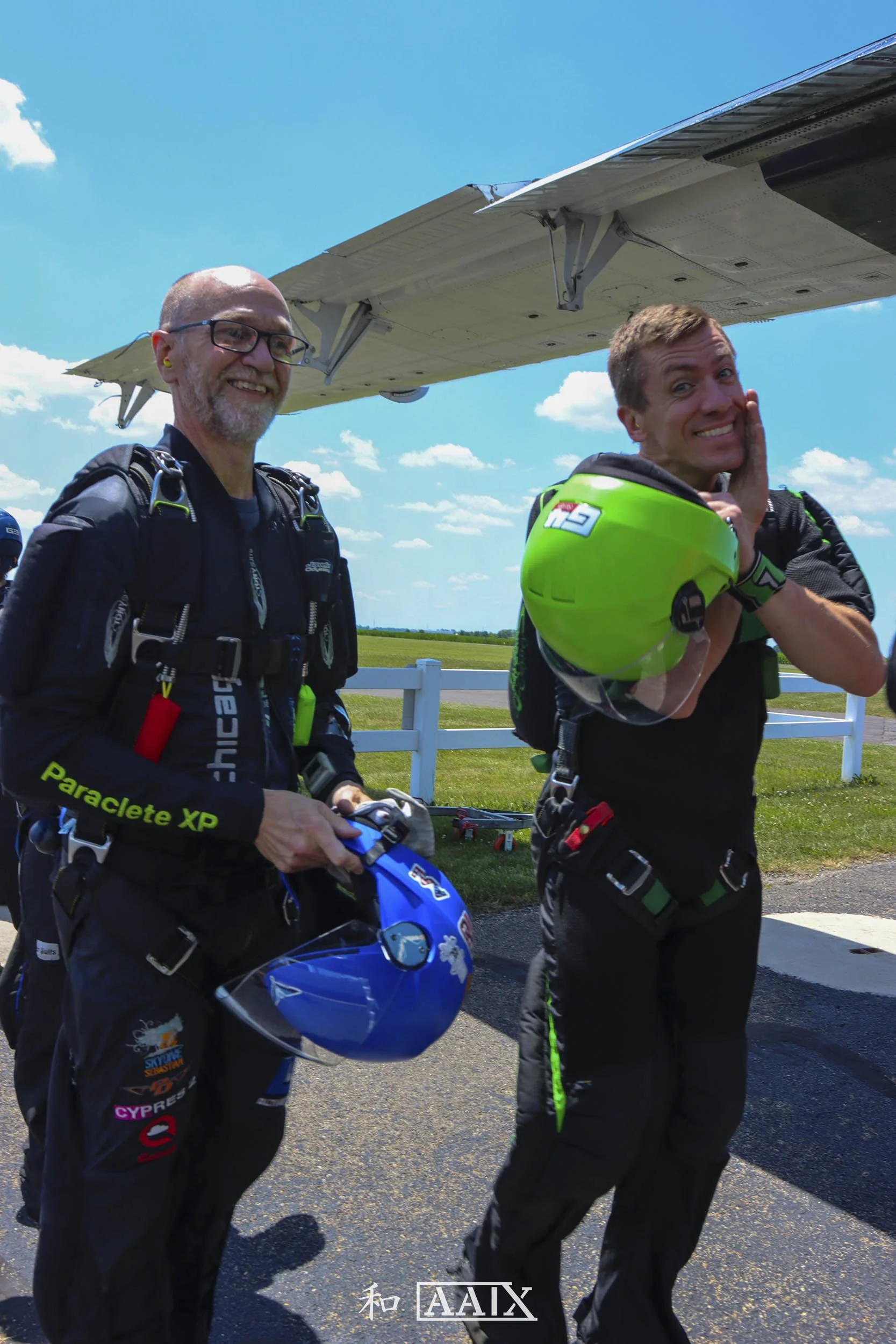 Two skydivers standing on the ground in front of a small airplane, both wearing jumpsuits and holding helmets, one smiling and the other with a smile on his face, under a bright blue sky with scattered clouds.