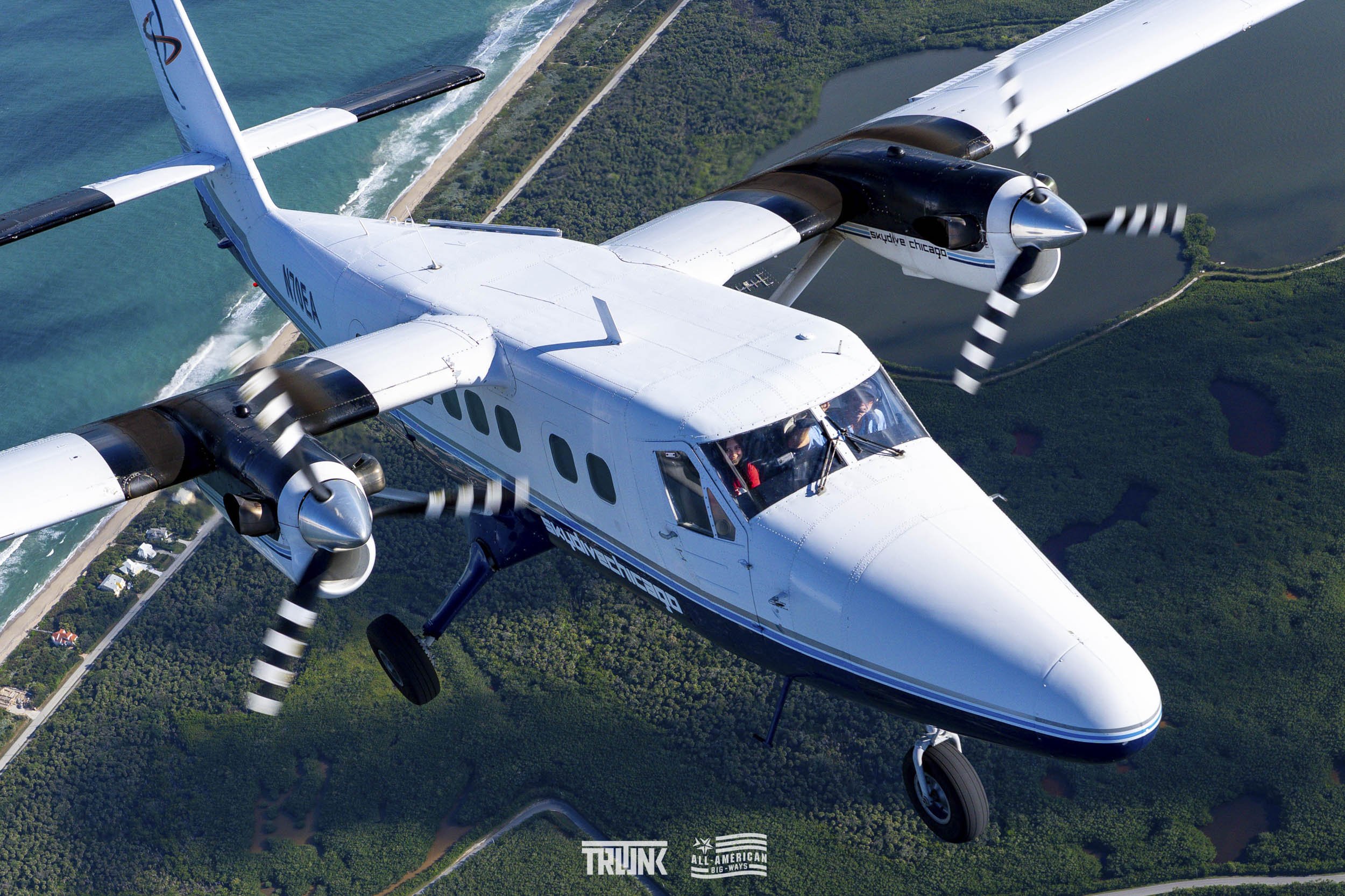 A twin-engine propeller airplane flying over a landscape of greenery, lakes, and a coastline.