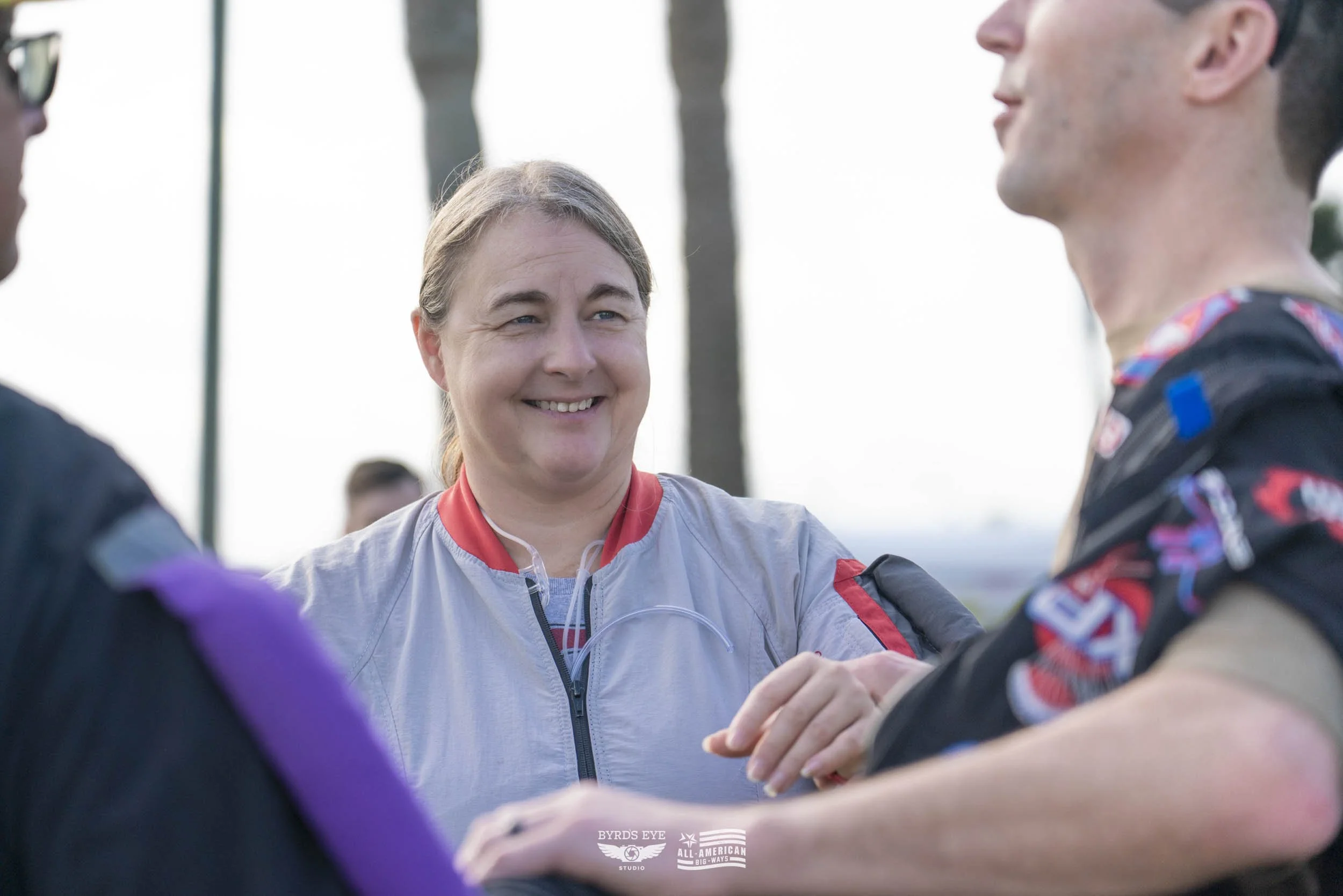 Three people outdoors, two men in racing suits shaking hands, woman smiling between them, palm trees in background.