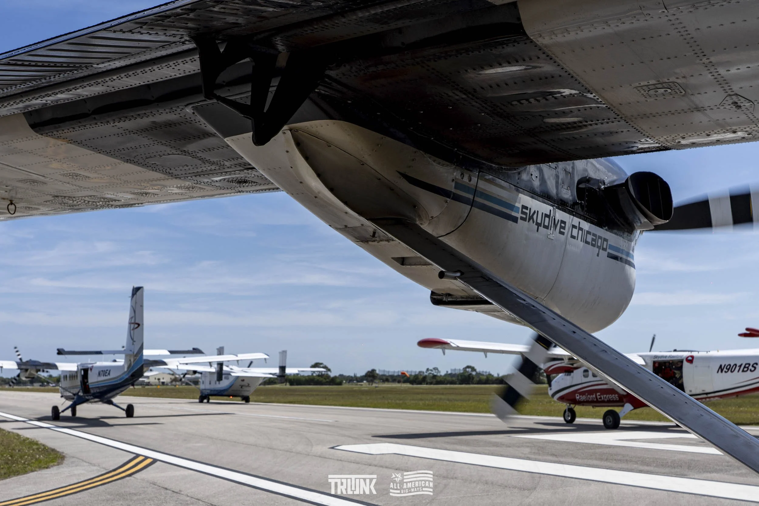 Close-up of a plane's wing and propeller with water decals, with several small planes parked on the tarmac and a blue sky in the background.