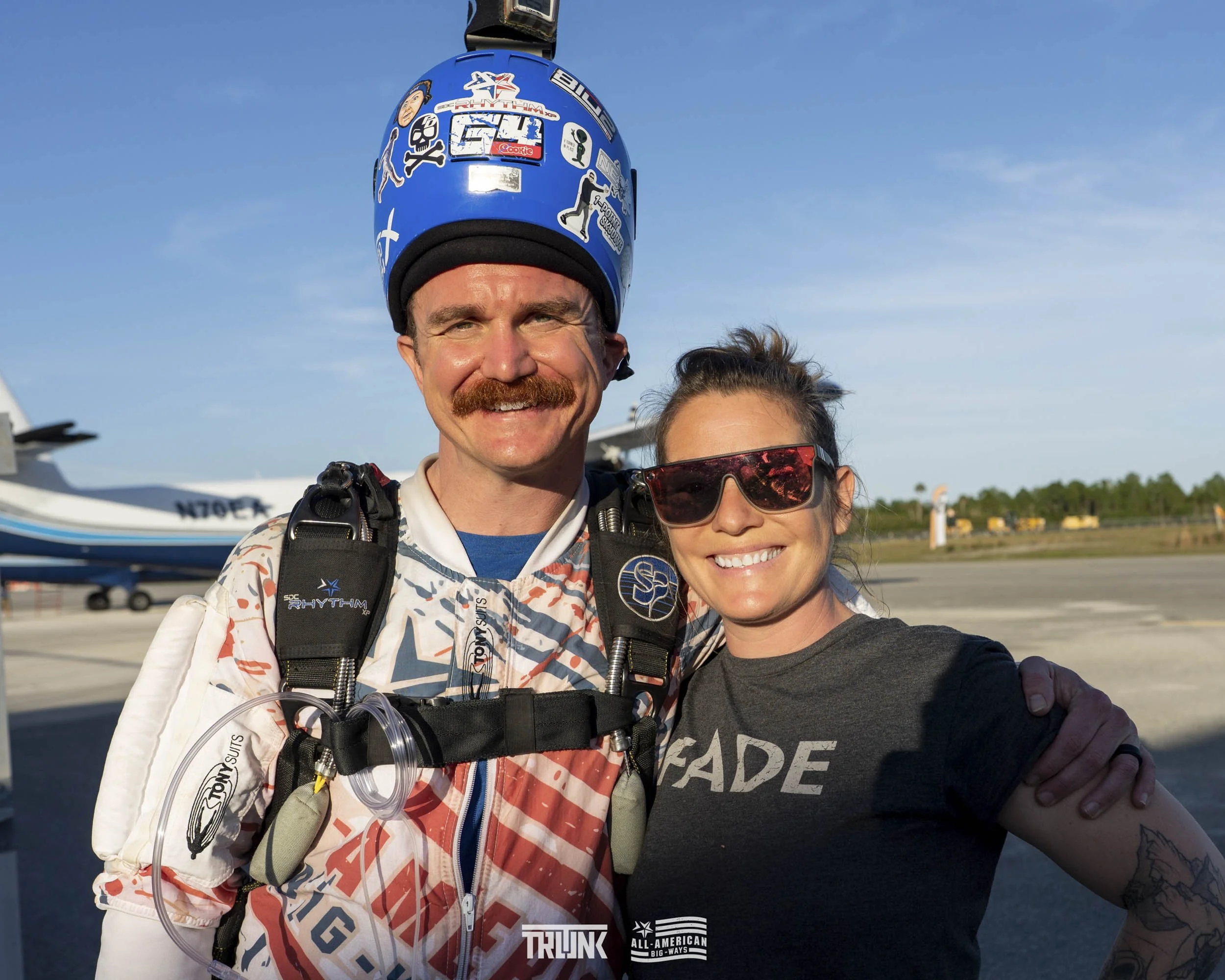 Two smiling skydivers, a man with a blue helmet and a woman with sunglasses, standing in an airport tarmac area with a small airplane in the background.
