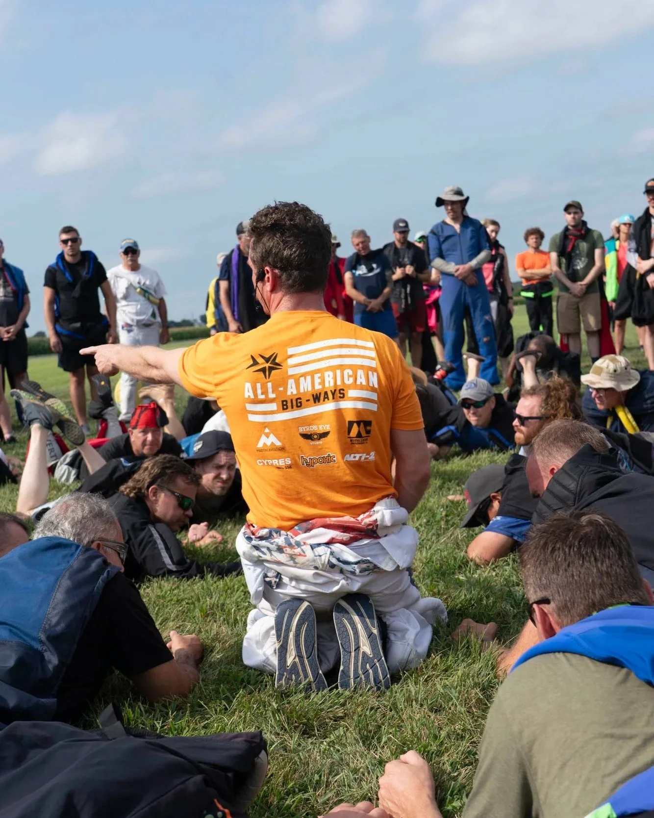 Group of people outdoors, some sitting and some standing, with a man in orange kneeling in the center giving a speech or instructions.