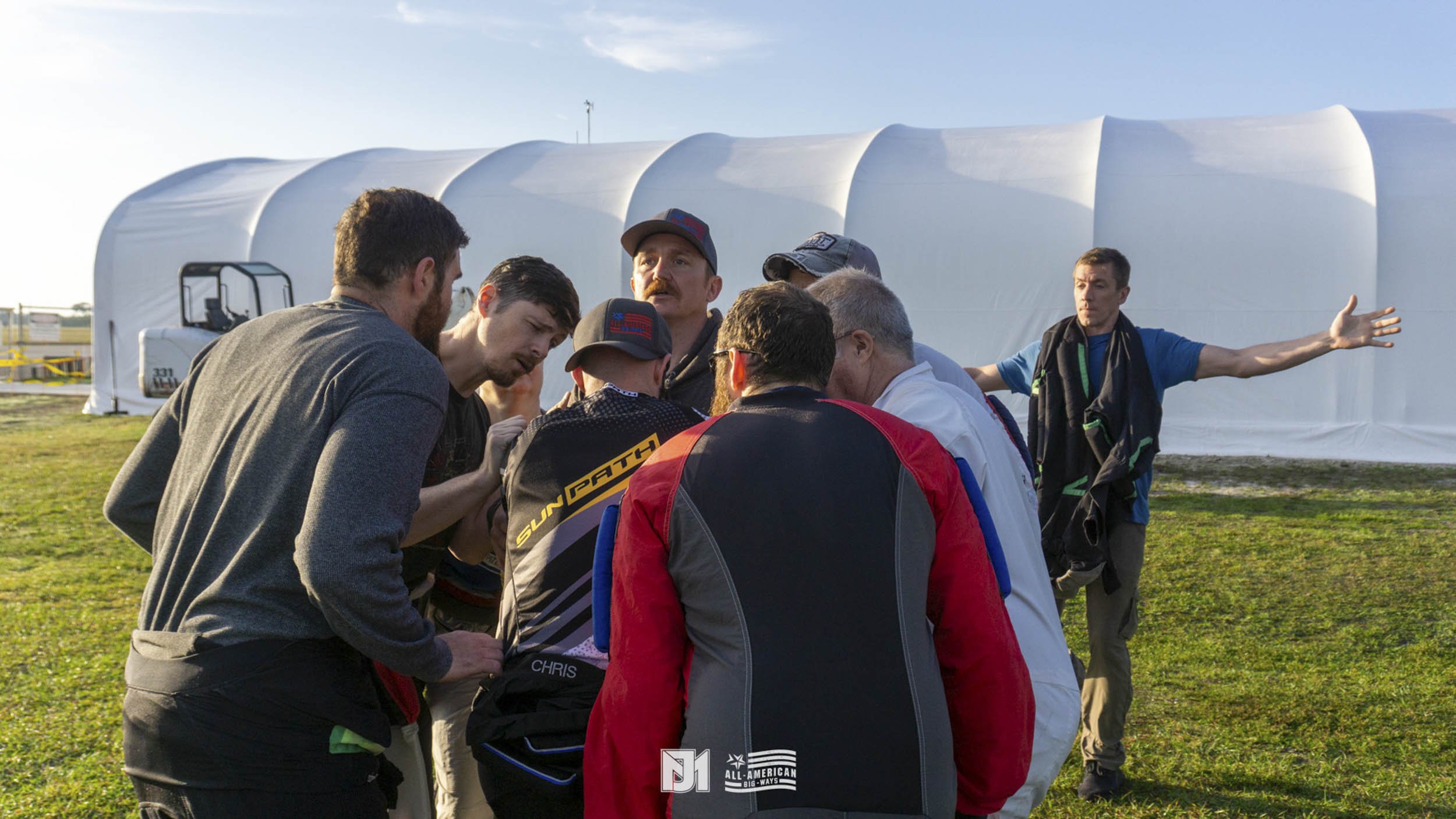 Group of people gathered outdoors near a large white tent, engaging in a discussion with one person gesturing.