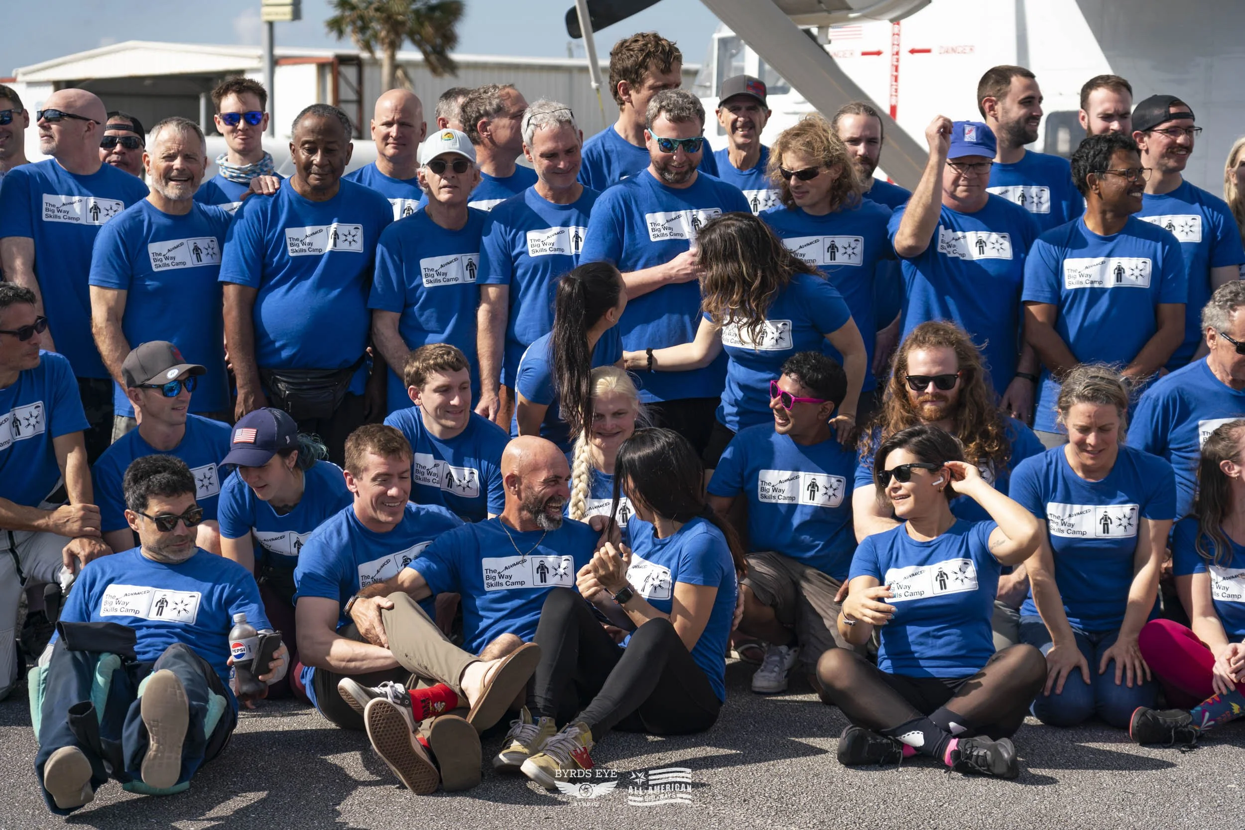 Group of people wearing matching blue t-shirts with a logo, gathered outdoors on a sunny day, some sitting on the ground and others standing, smiling and socializing.