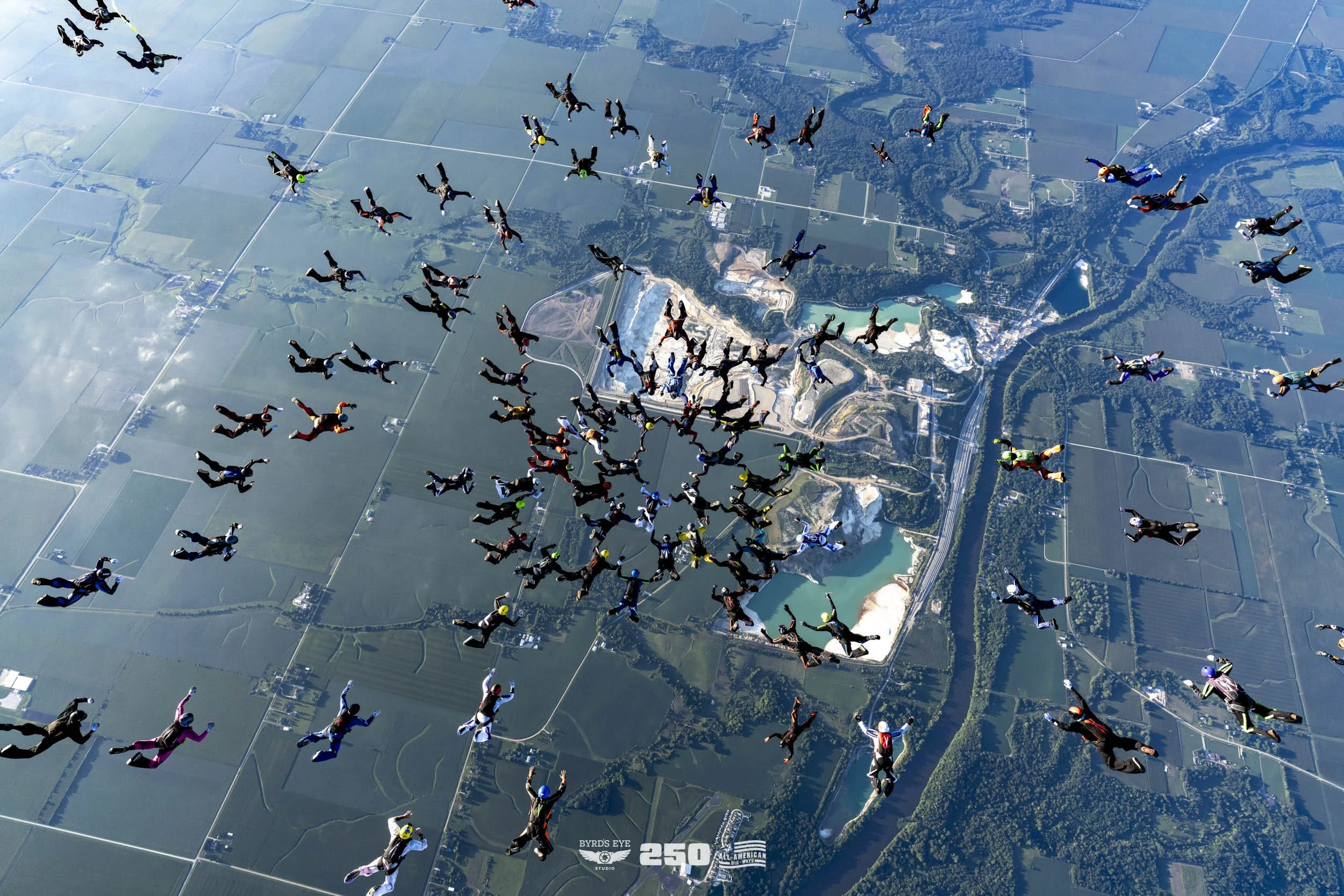 Group of skydivers in free fall over a landscape with lakes, forests, and farmland.