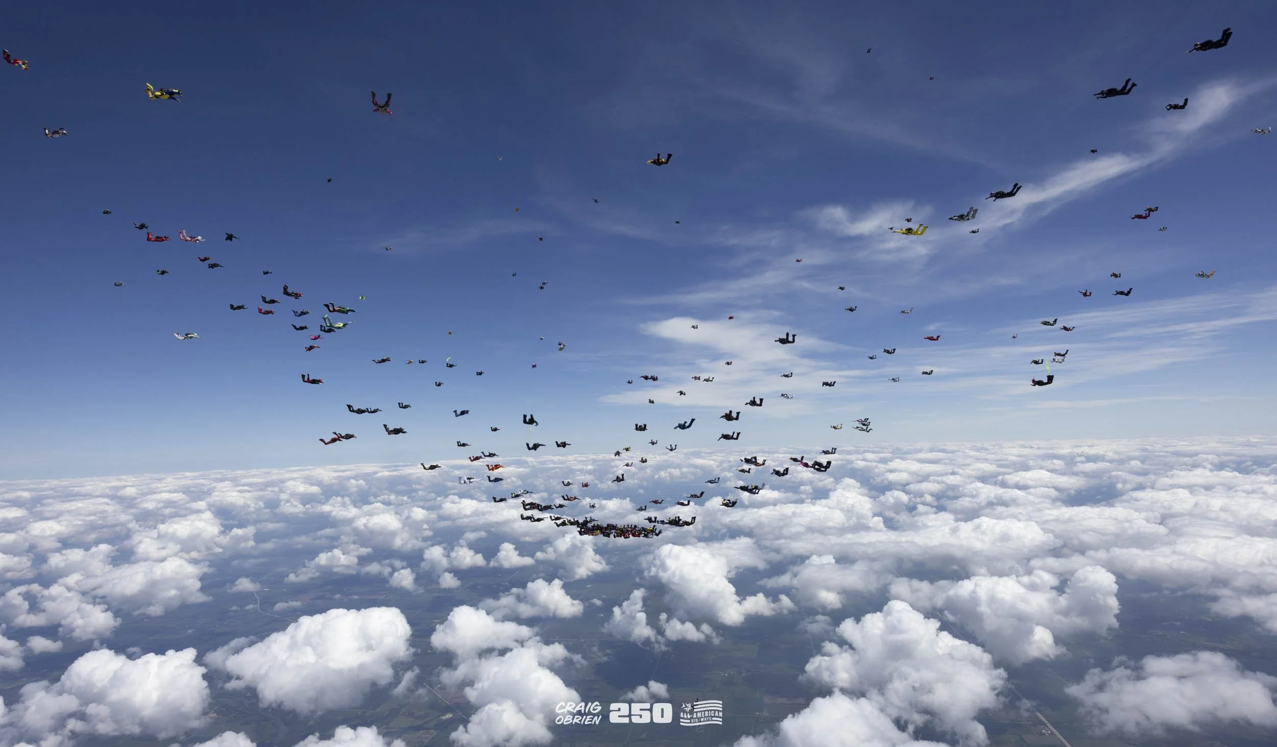 Skydivers in free fall above a cloud-filled landscape with a blue sky and scattered clouds, celebrating for Craig O'Brien's 250th dive anniversary.