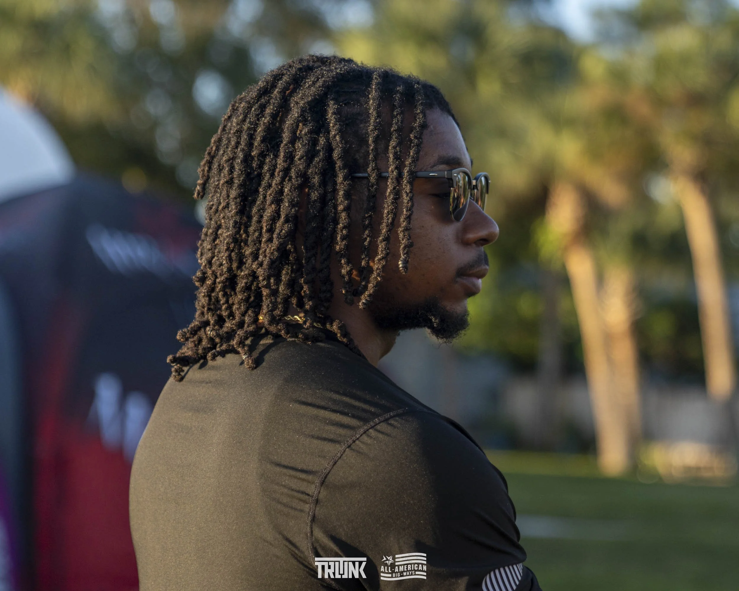 Side profile of a young man with dreadlocks wearing sunglasses outdoors, with trees and a grassy area in the background.
