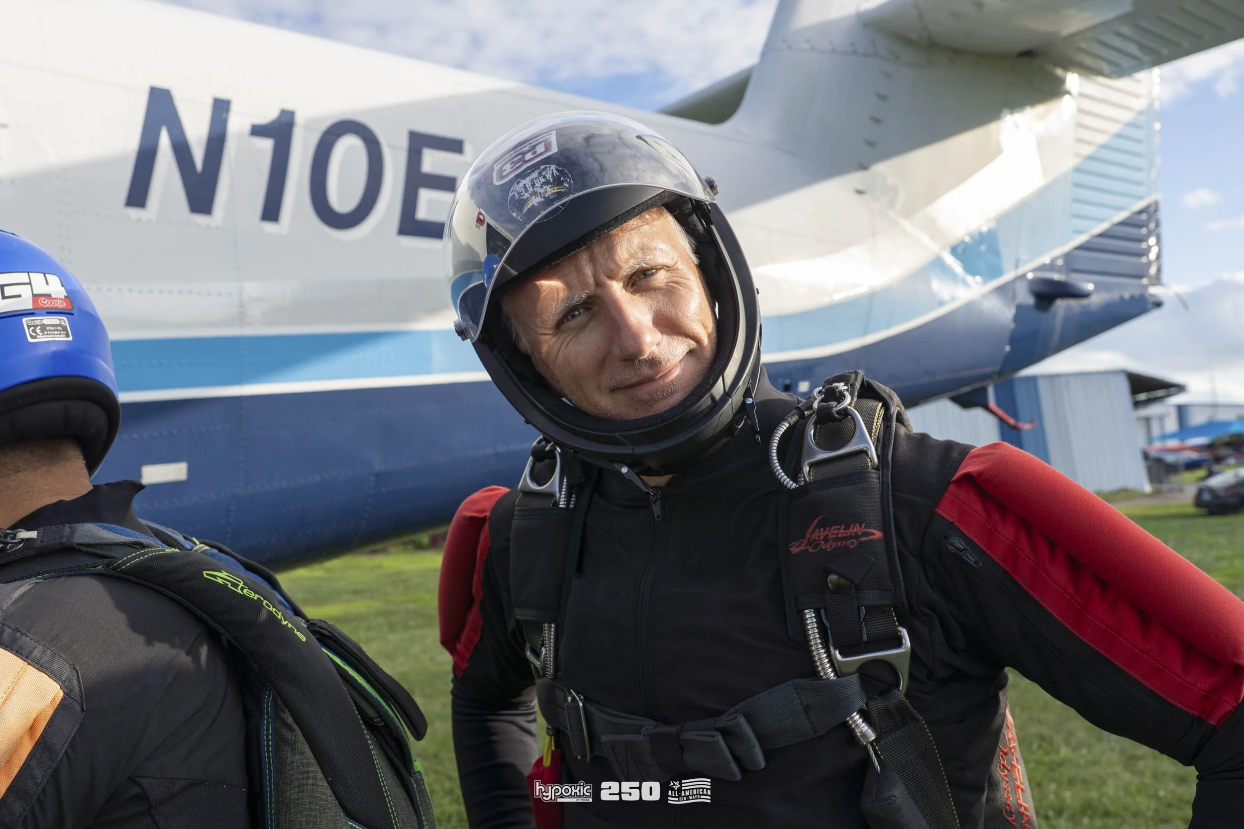 A man in skydiving gear, including a helmet, standing in front of an airplane on a grassy field.