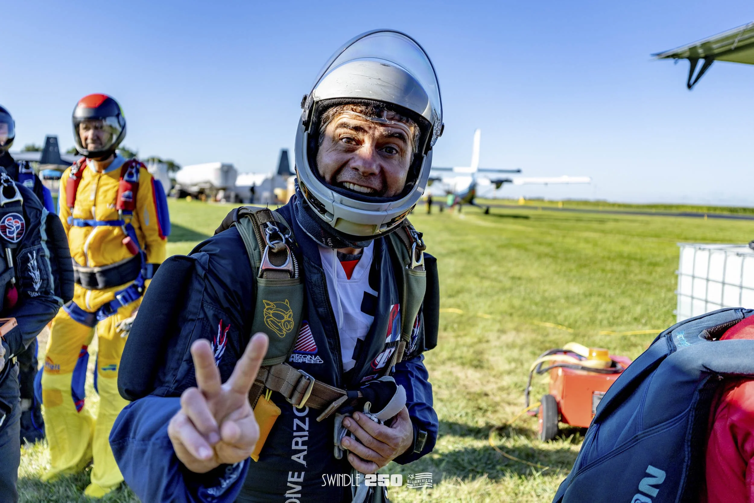 Excited man in a helmet and skydiving gear making a peace sign at an airfield, with other skydivers and planes in the background.