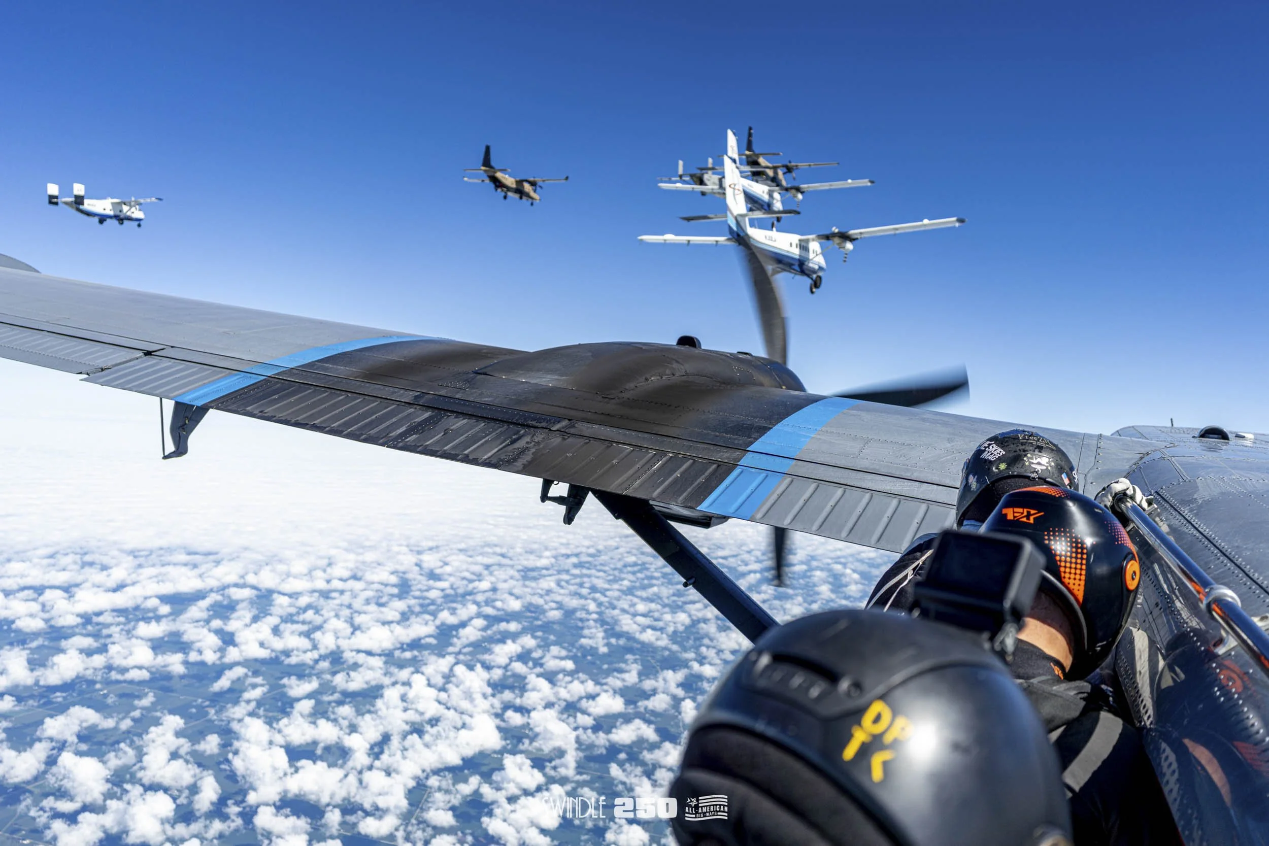 In-flight view from a drone or aircraft showing a person in a cockpit with a helmet, flying above clouds through a clear blue sky, with several military aircraft flying in formation nearby.