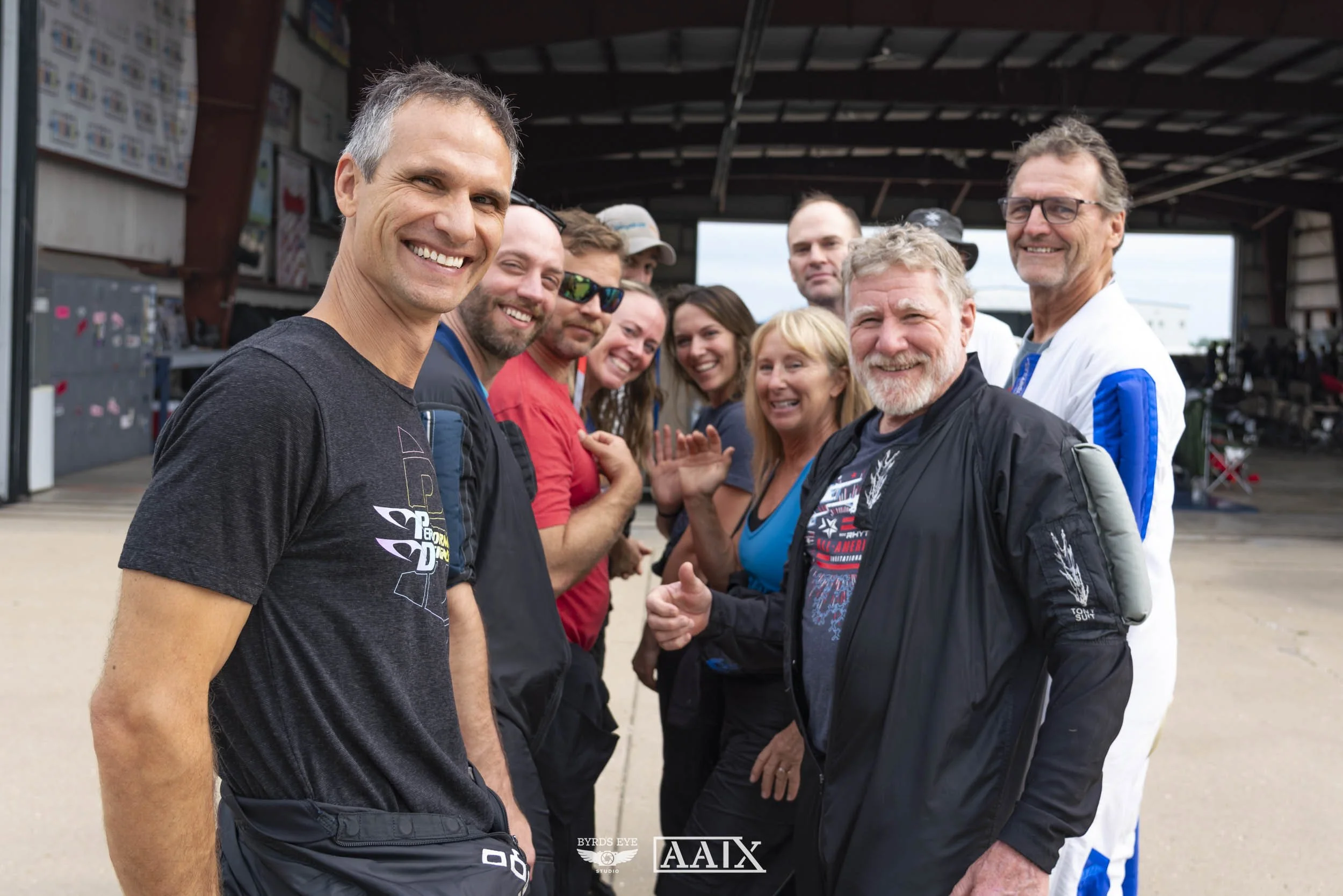 A group of smiling people standing in a hangar or open-air structure at an airfield, with some wearing casual clothing and others in uniforms, possibly pilots or crew members.