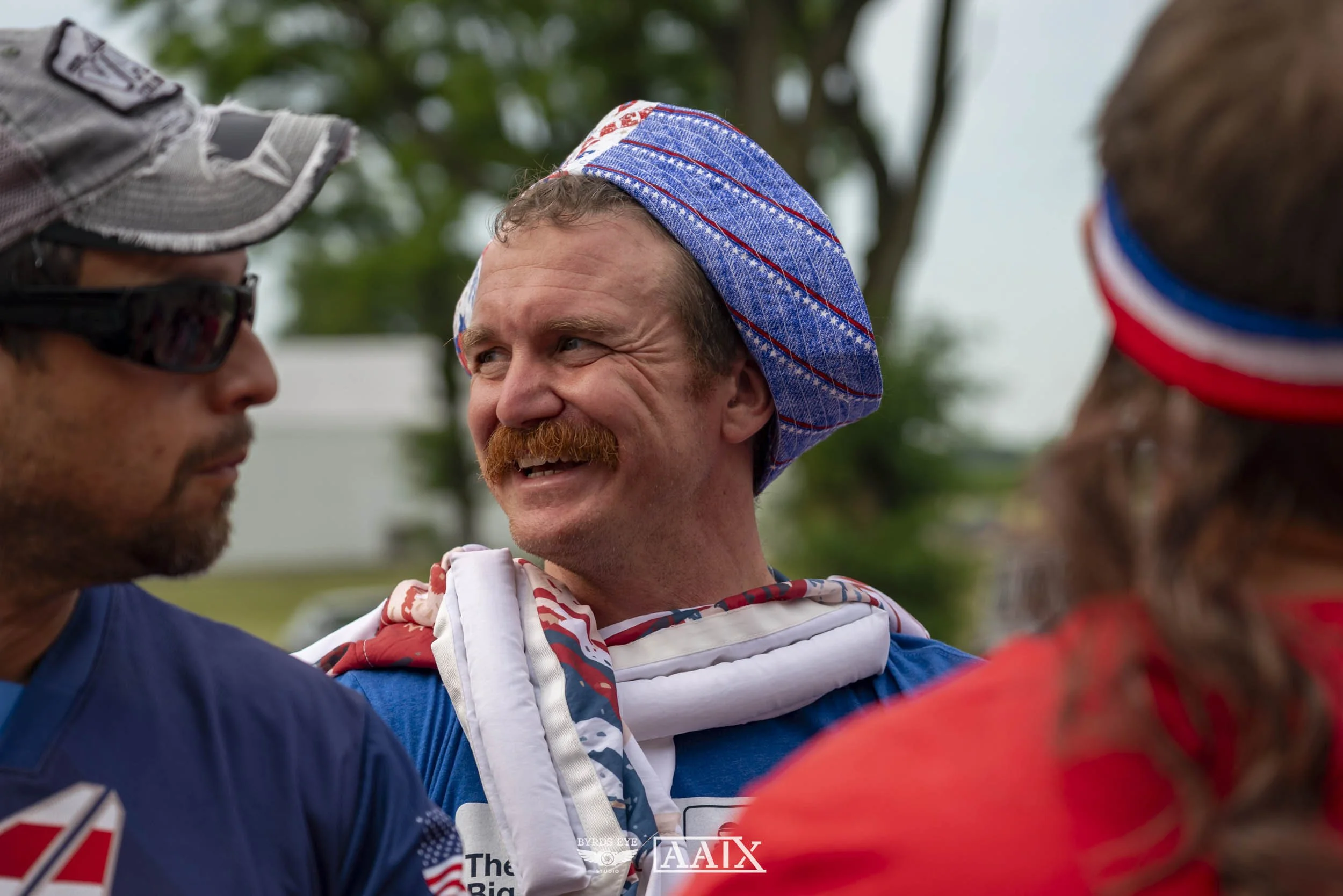 A man with a red mustache and beard, wearing a blue cap and cheering with other people outdoors.