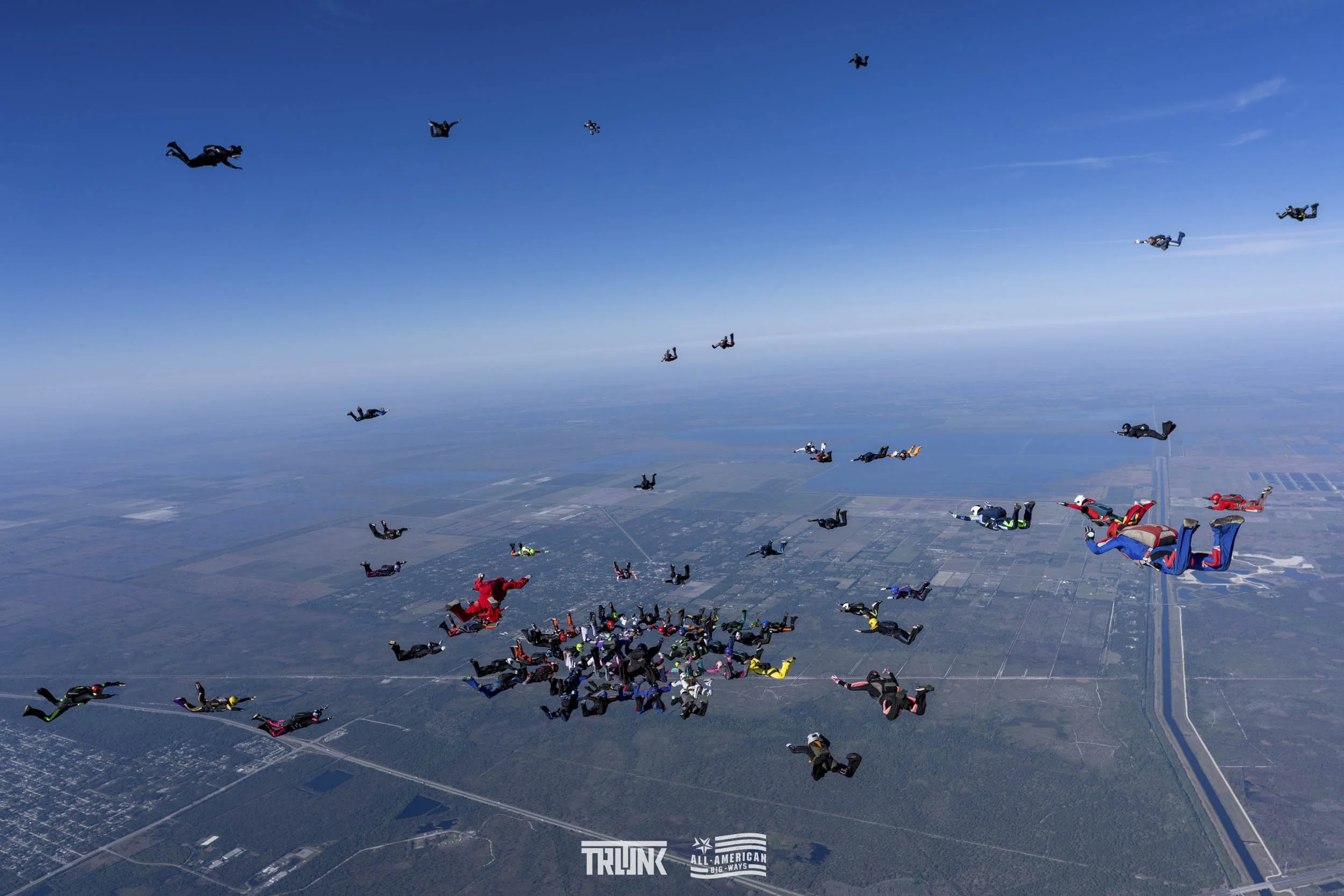Numerous skydivers in free fall over a landscape, some with colorful jumpsuits, forming a group with a clear sky.
