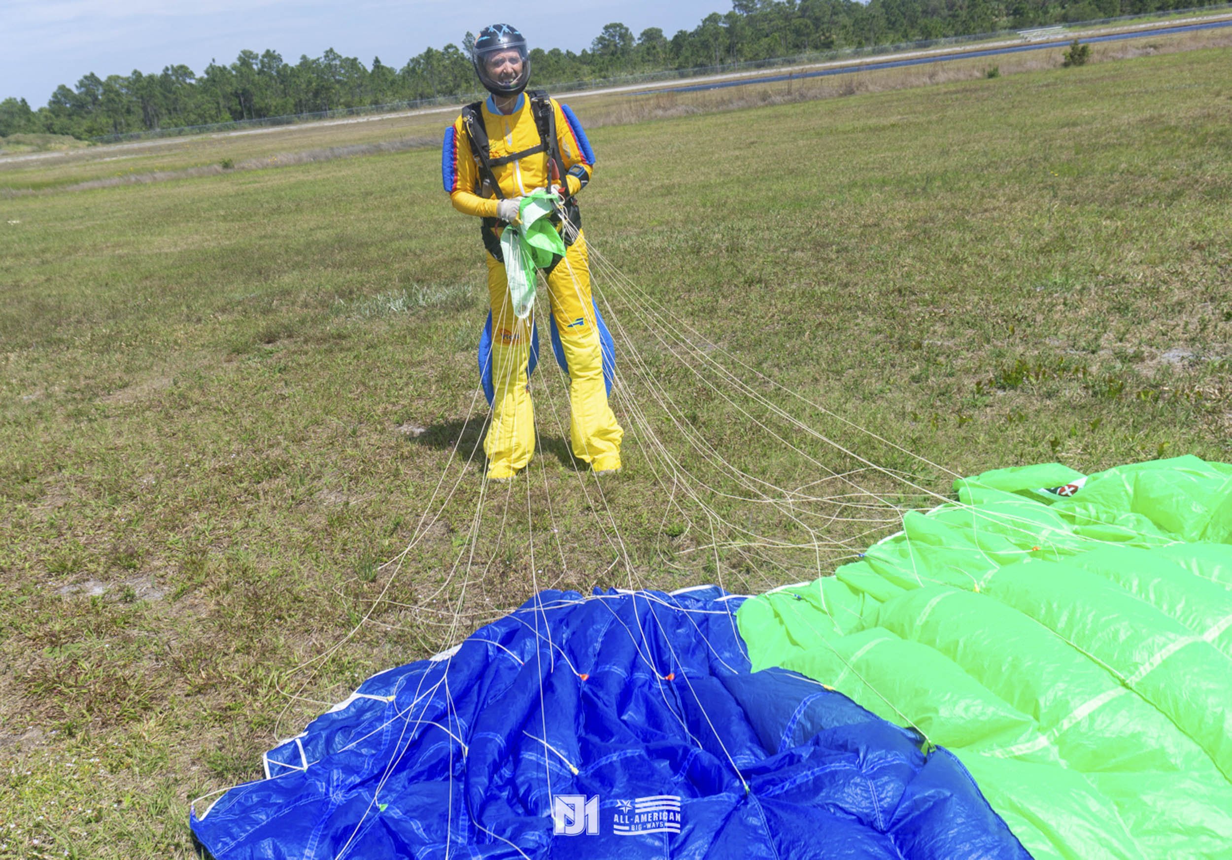 A person preparing for paragliding on a grassy field, wearing a helmet, yellow and blue gear, and holding a green and white parachute.