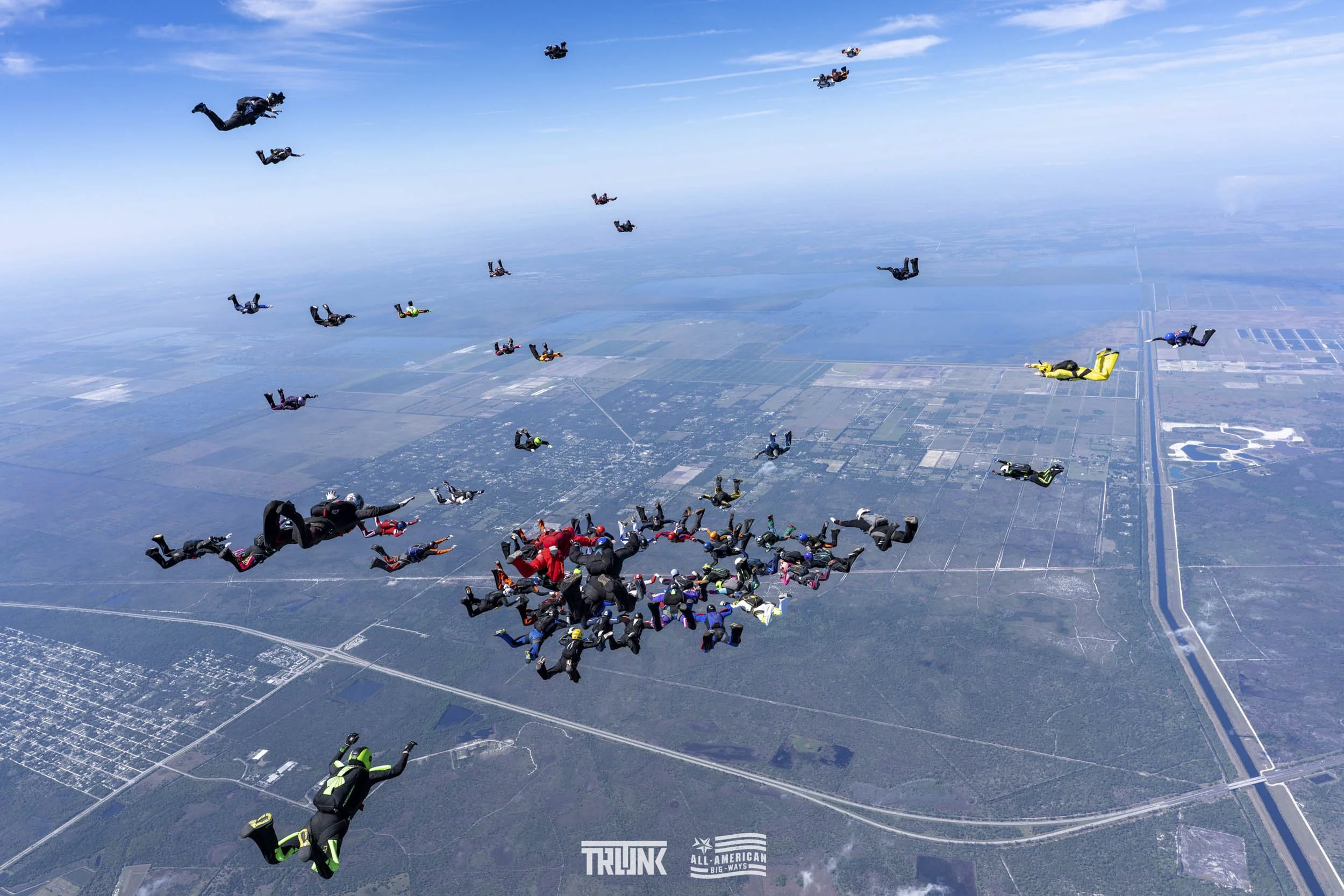 A large group of skydivers free-falling in the sky over a landscape with fields and roads below.