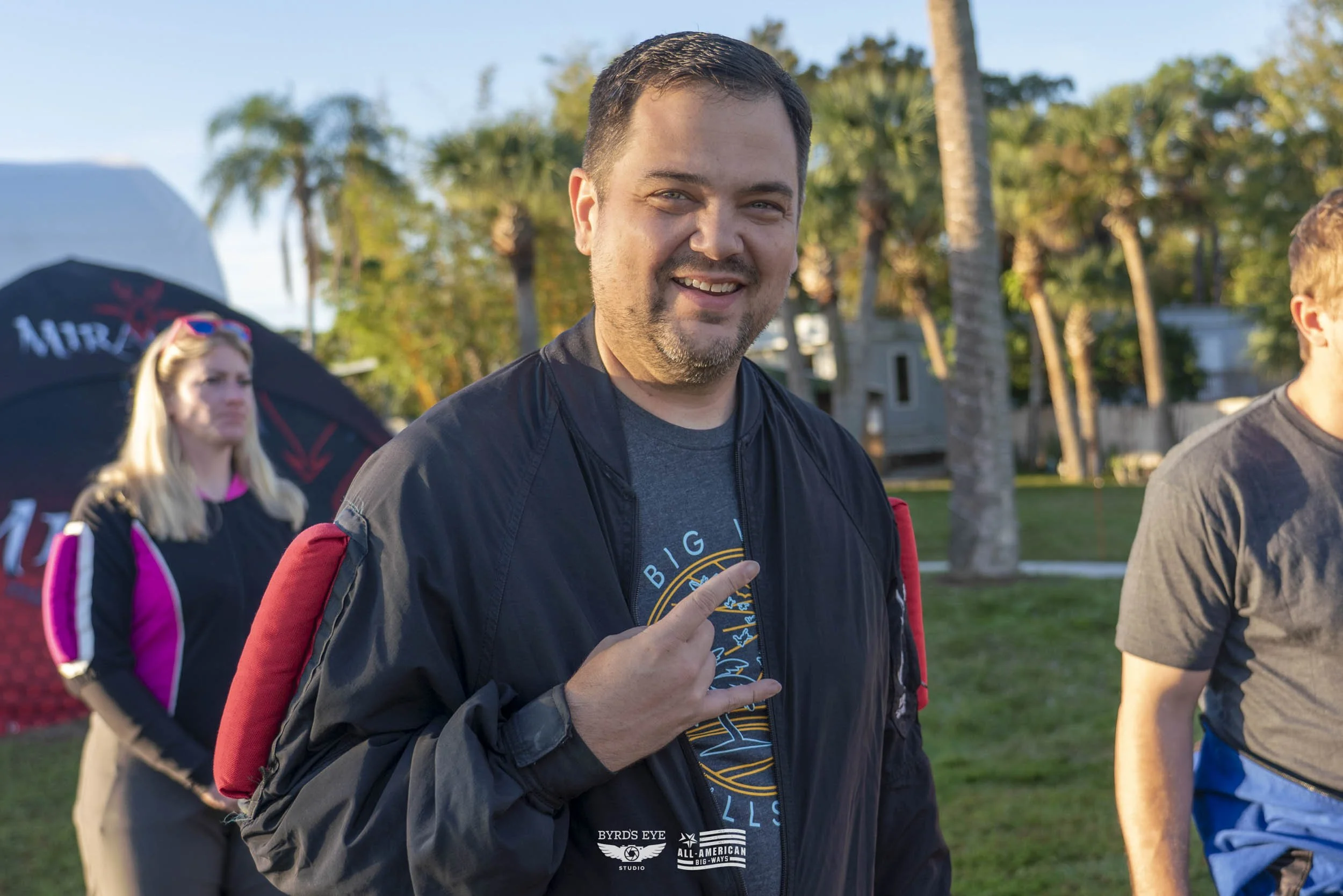 Smiling man making a rock and roll hand gesture at an outdoor event, with palm trees and other people in the background.